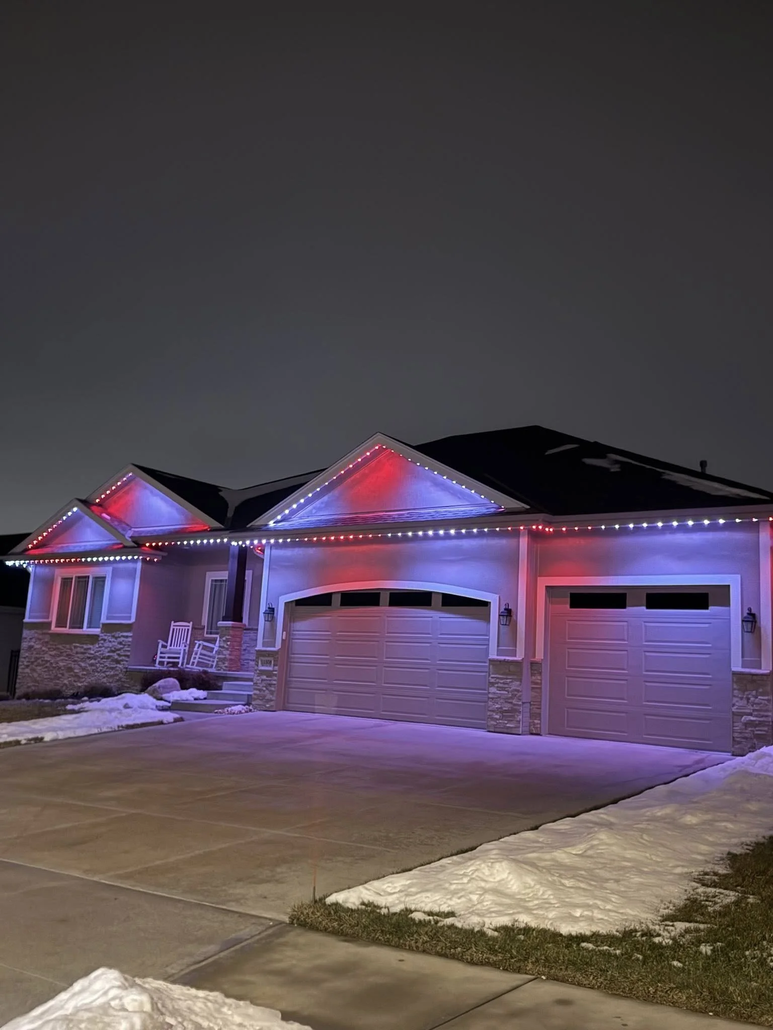 A house with a driveway at night decorated with red, white, and blue string lights on the roofline.