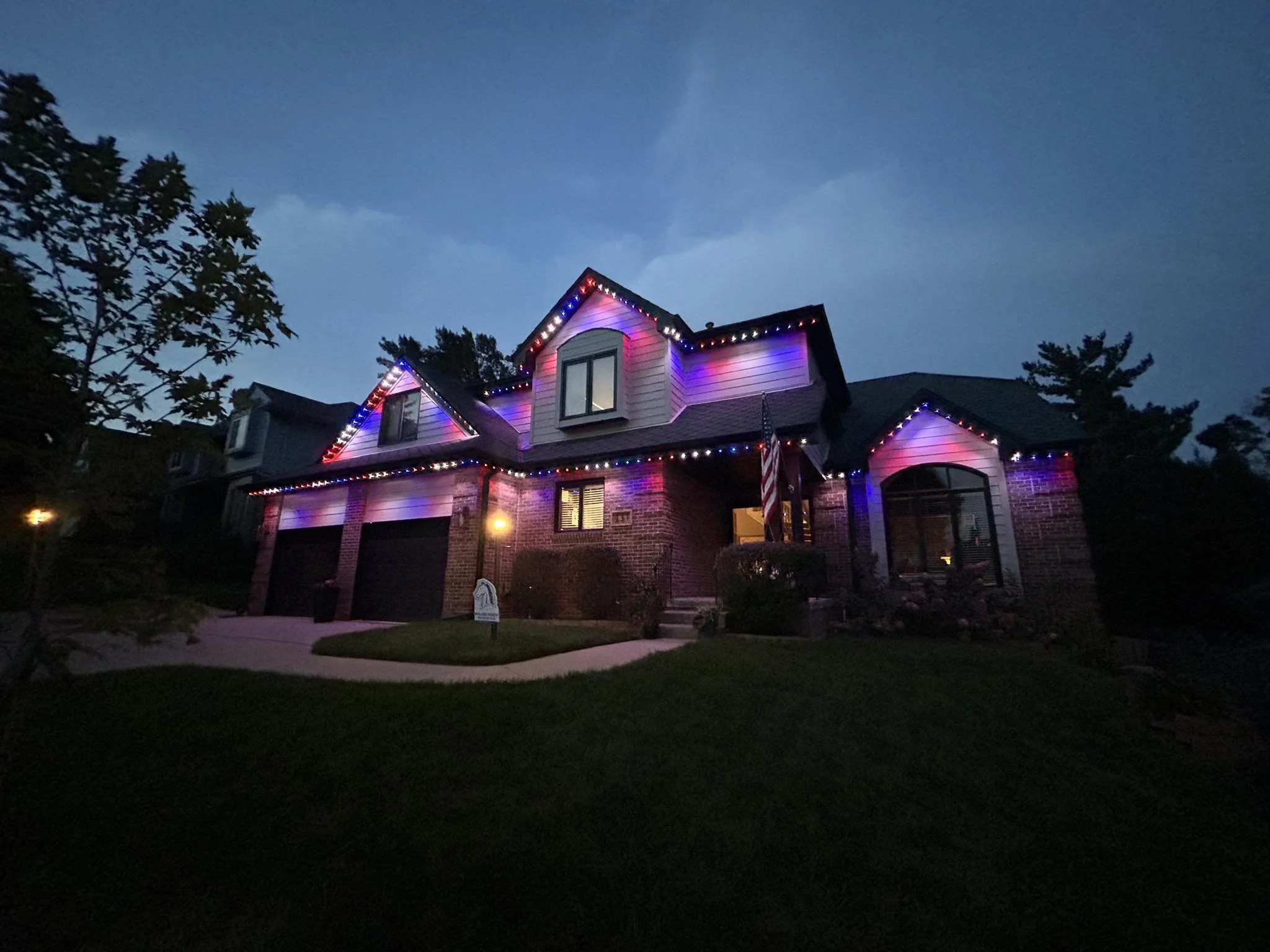 A two-story house at night decorated with multicolored string lights along the roofline, with a manicured lawn and a sidewalk in front.