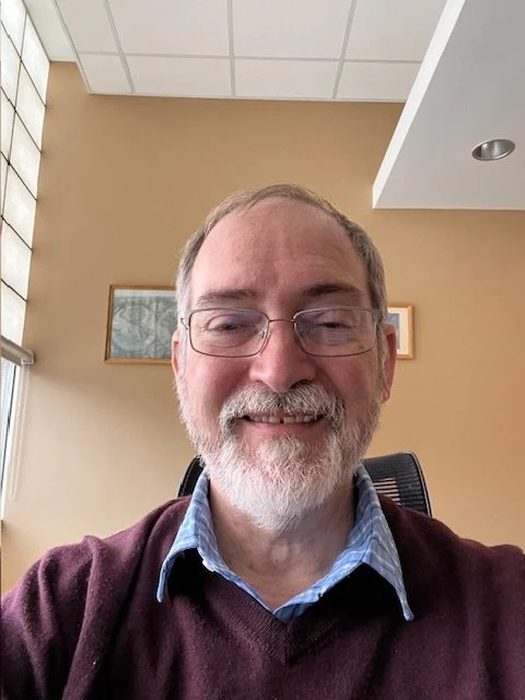 A middle-aged man with glasses, a gray beard, and short gray hair, smiling at the camera, sitting in an office with beige walls, framed pictures, and a window with blinds.