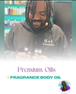 A man with dreadlocks and a beard smiling in a store aisle holding essential oils, with a sign reading 'Premium Oils' and 'Fragrance Body Oil' in the background.