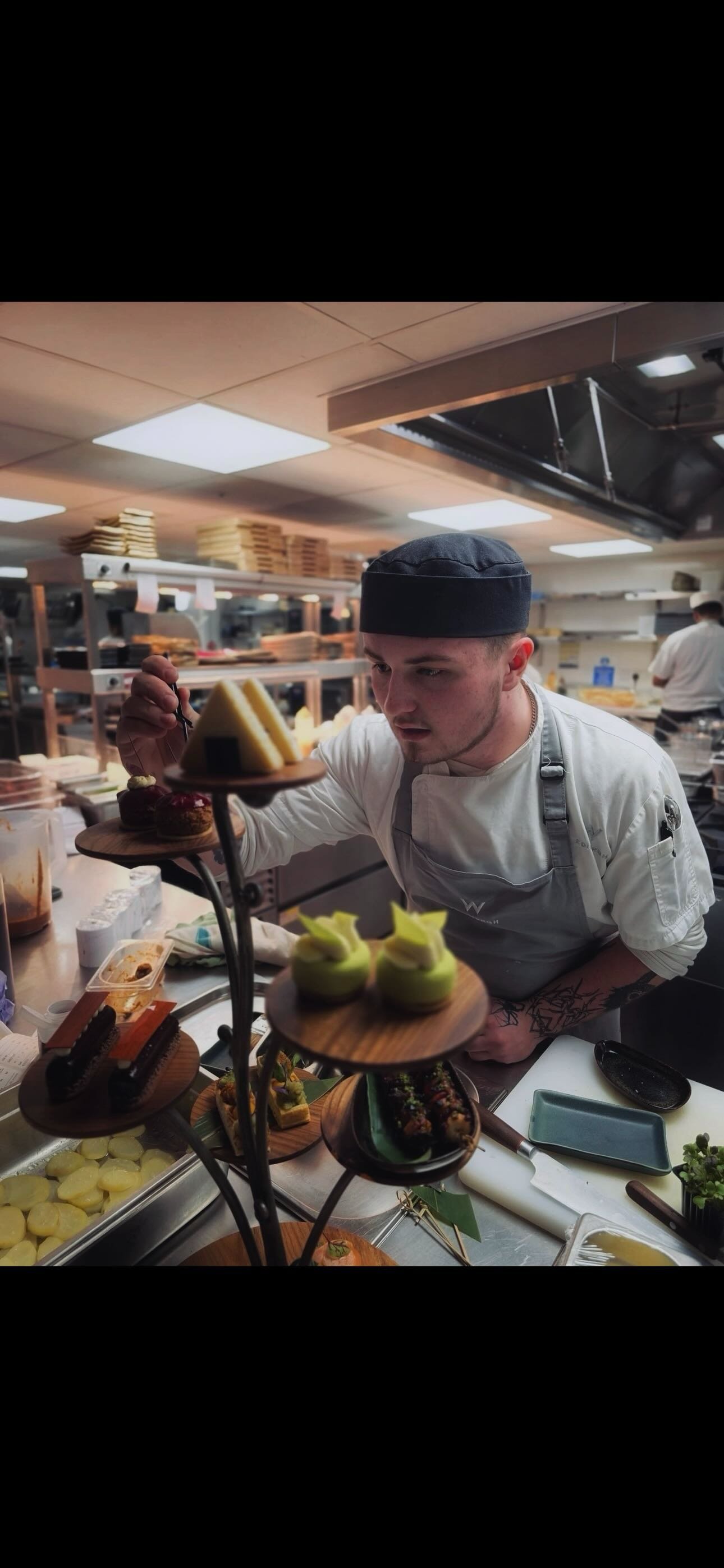 A male chef with a black hat and gray apron arranging desserts on a display stand in a professional kitchen.