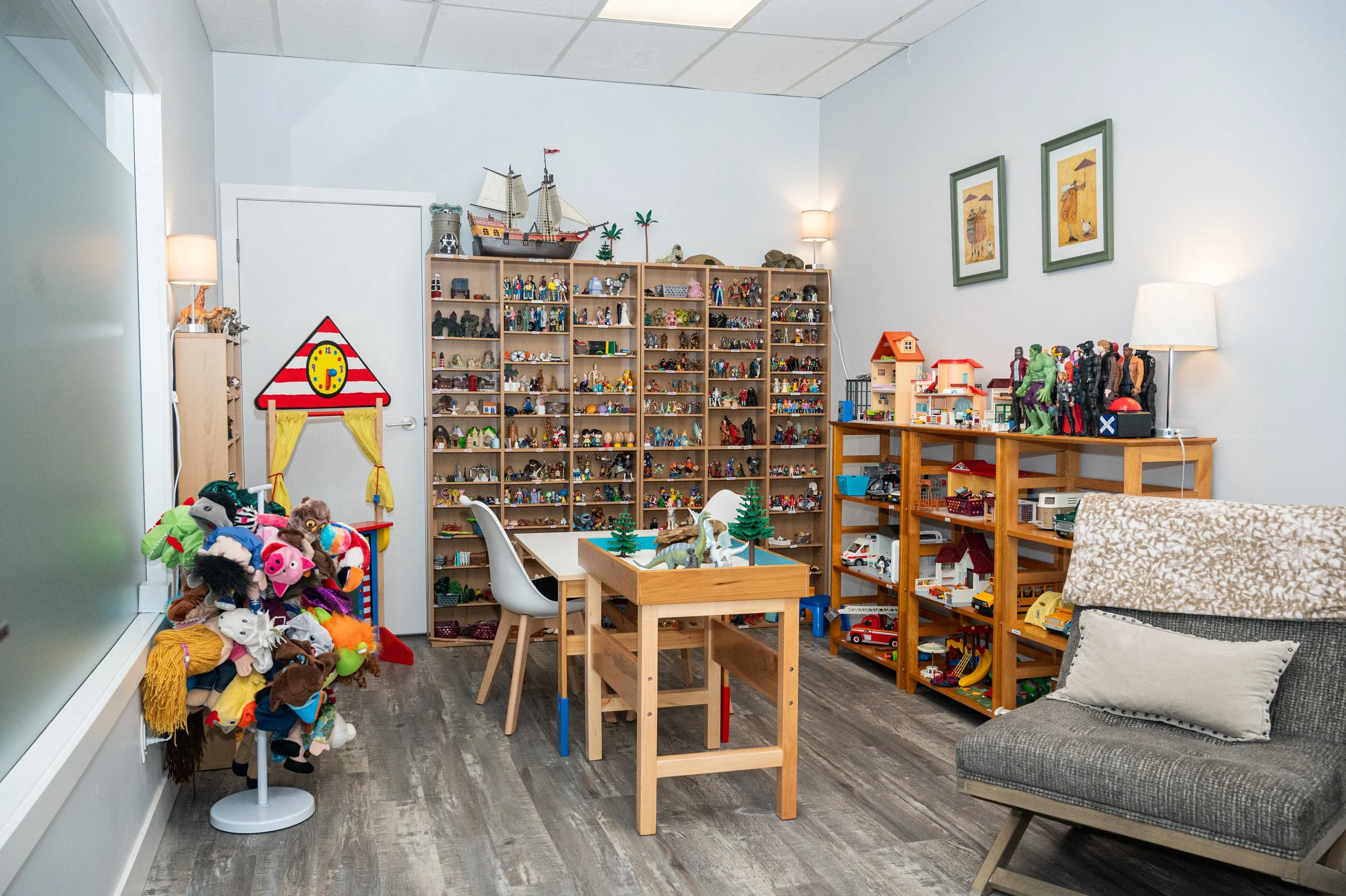 A room filled with various toys, shelves, and furniture. There's a table with some dinosaur figurines, a chair, a gray couch, and a display of numerous small figurines and toys on shelves and racks.