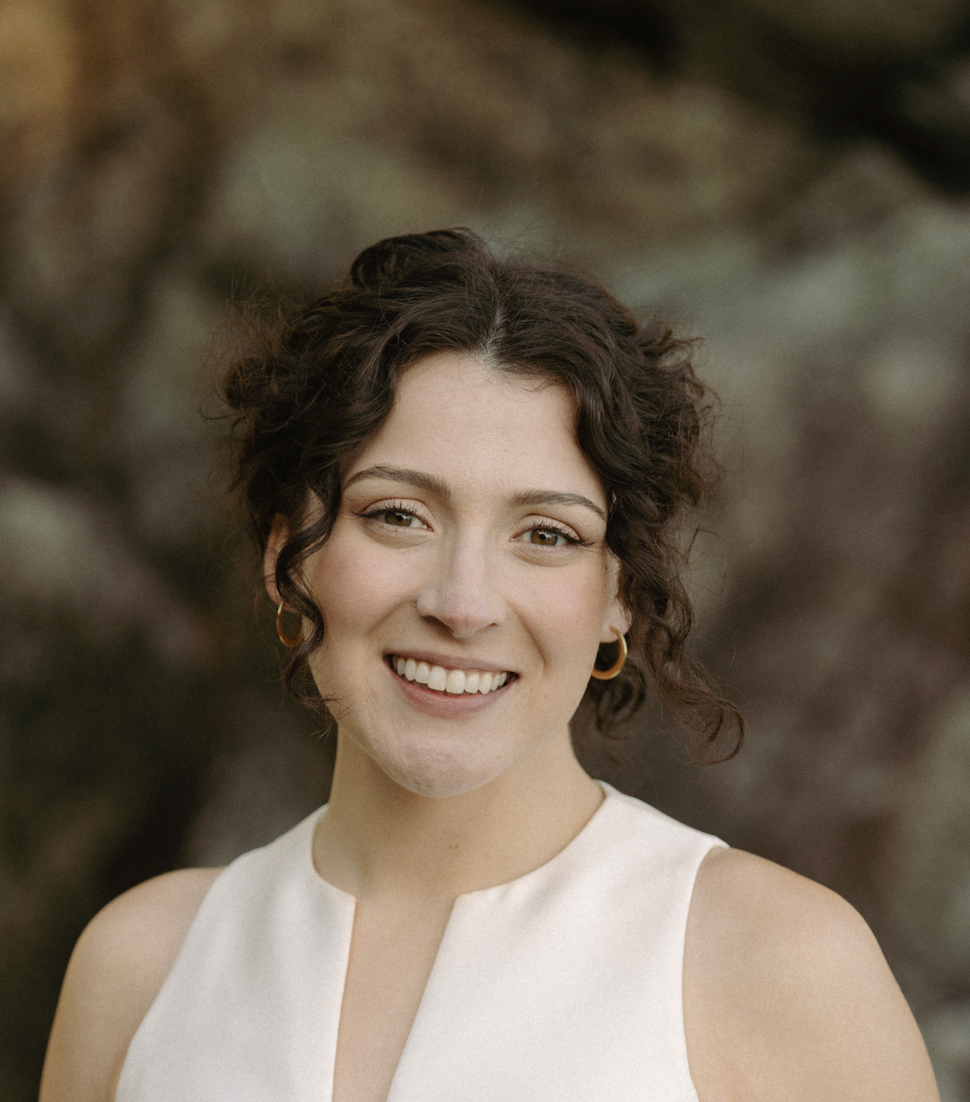A smiling woman with curly dark hair and light makeup, wearing a sleeveless white top and gold hoop earrings, outdoors with a blurred natural background.