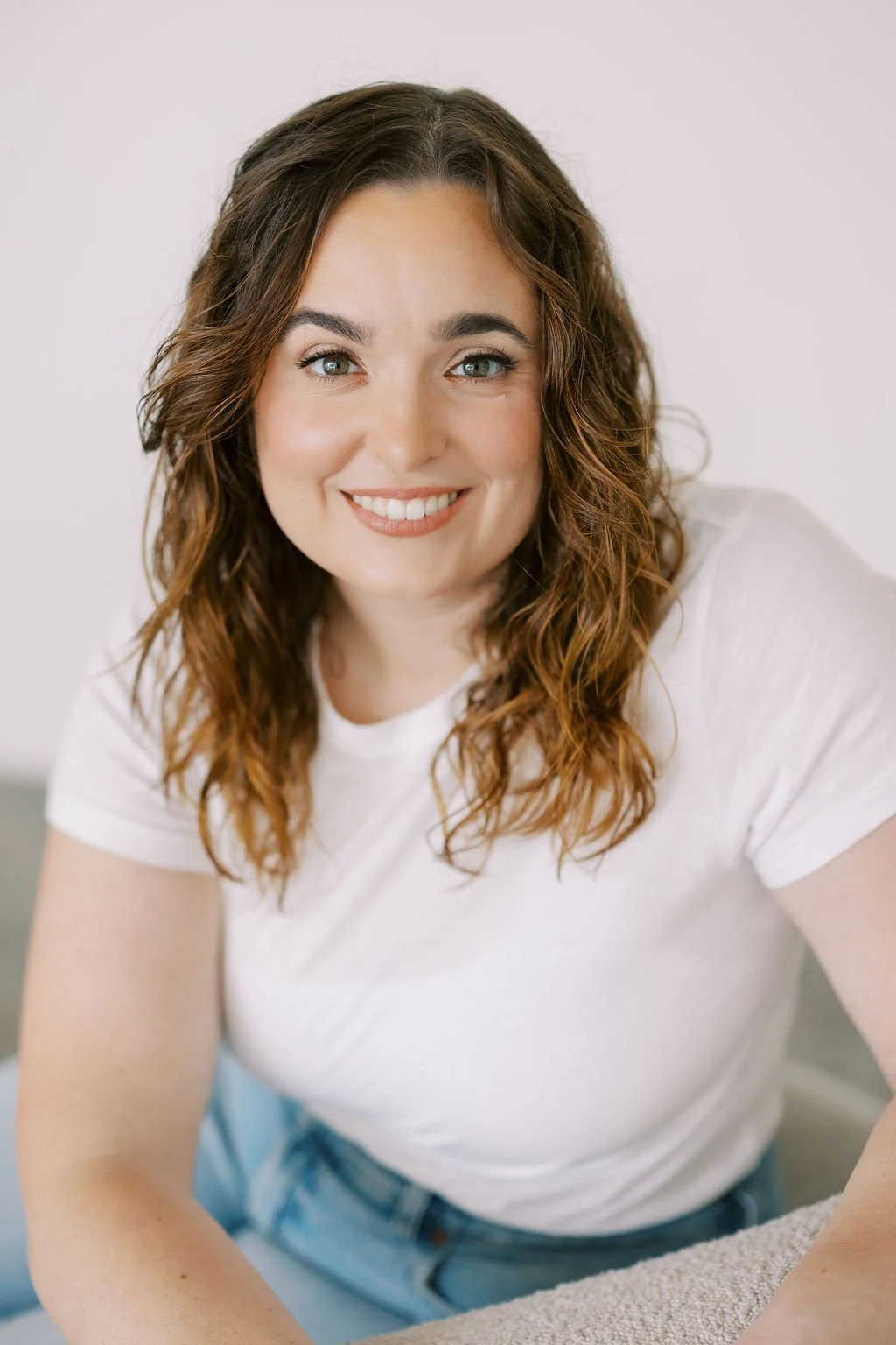 A woman with curly brown hair and blue eyes smiling, wearing a white t-shirt, sitting on a grey couch in a bright room.