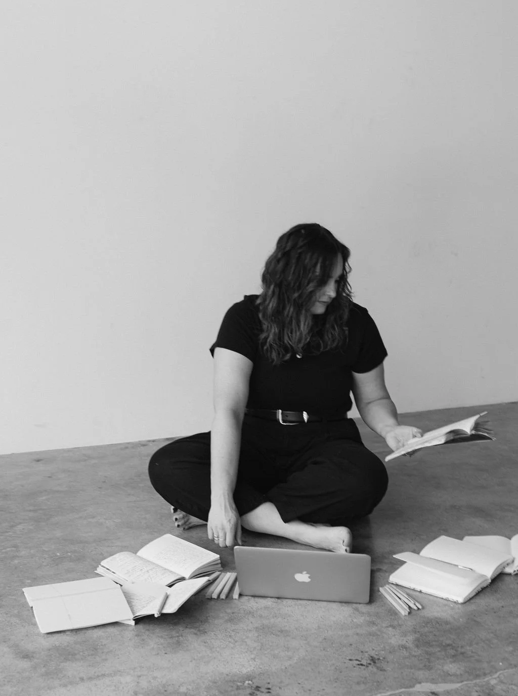 Woman sitting on the floor surrounded by open books, a laptop, and notebooks, reading a book in a minimalistic room.