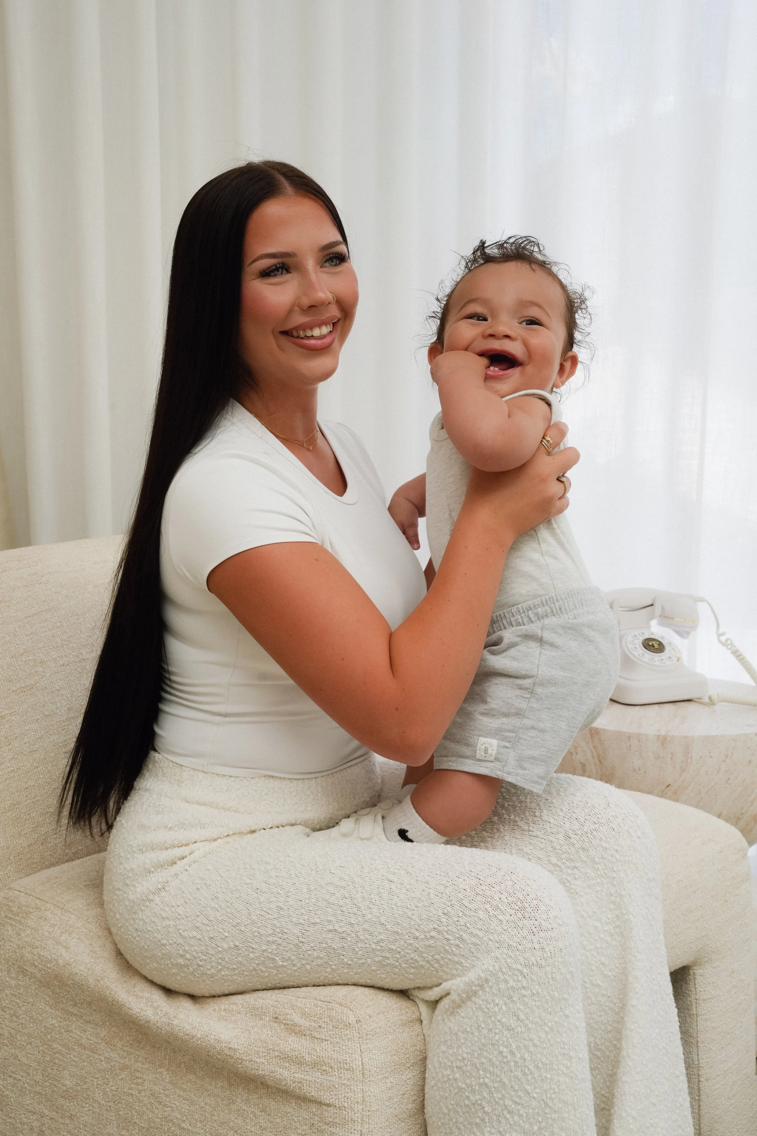 A woman with long black hair smiling while holding a laughing baby boy dressed in light gray clothing, sitting on a beige sofa with a cream-colored backdrop.