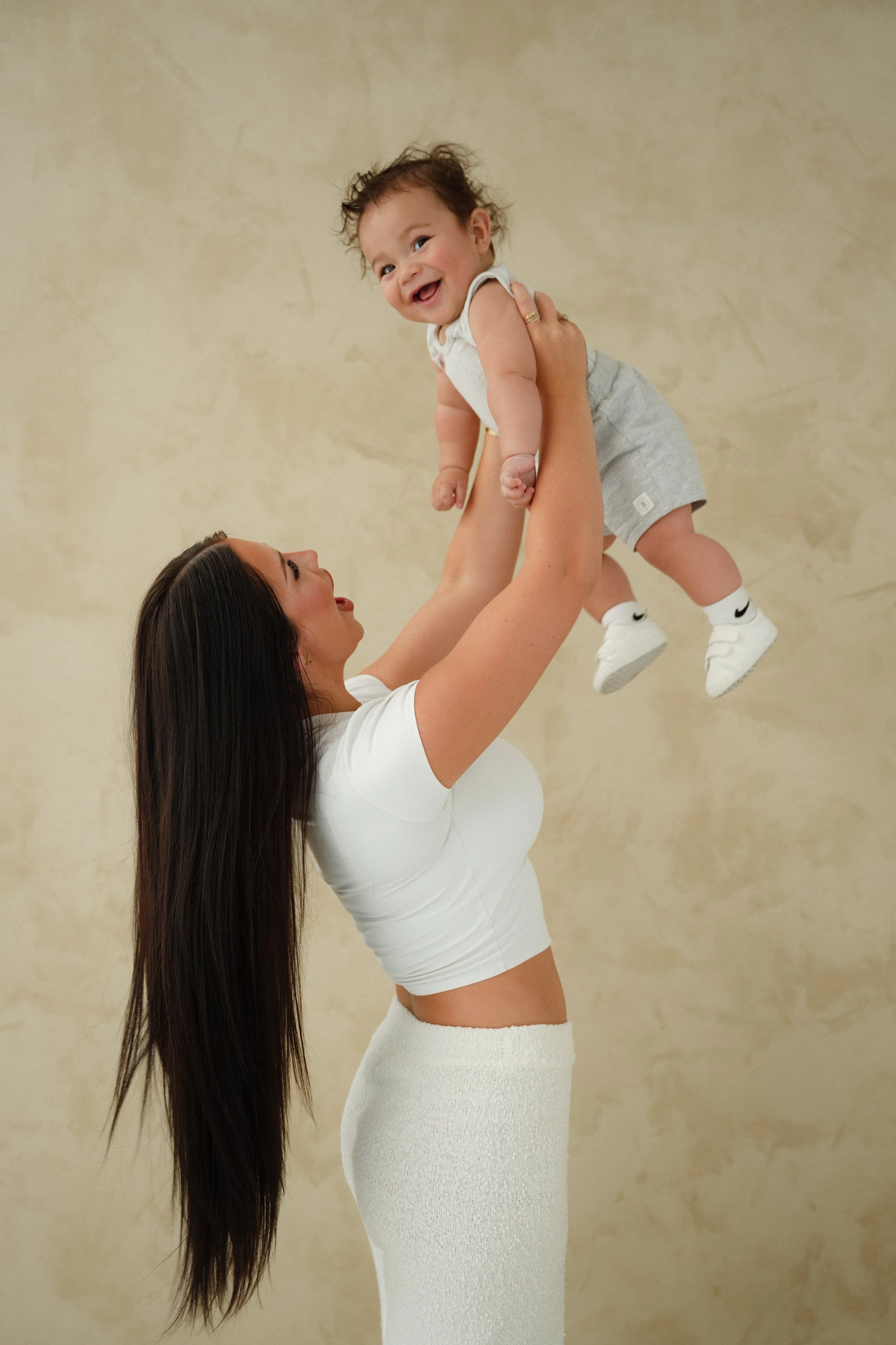 A woman lifting a happy baby with messy hair in a beige background.
