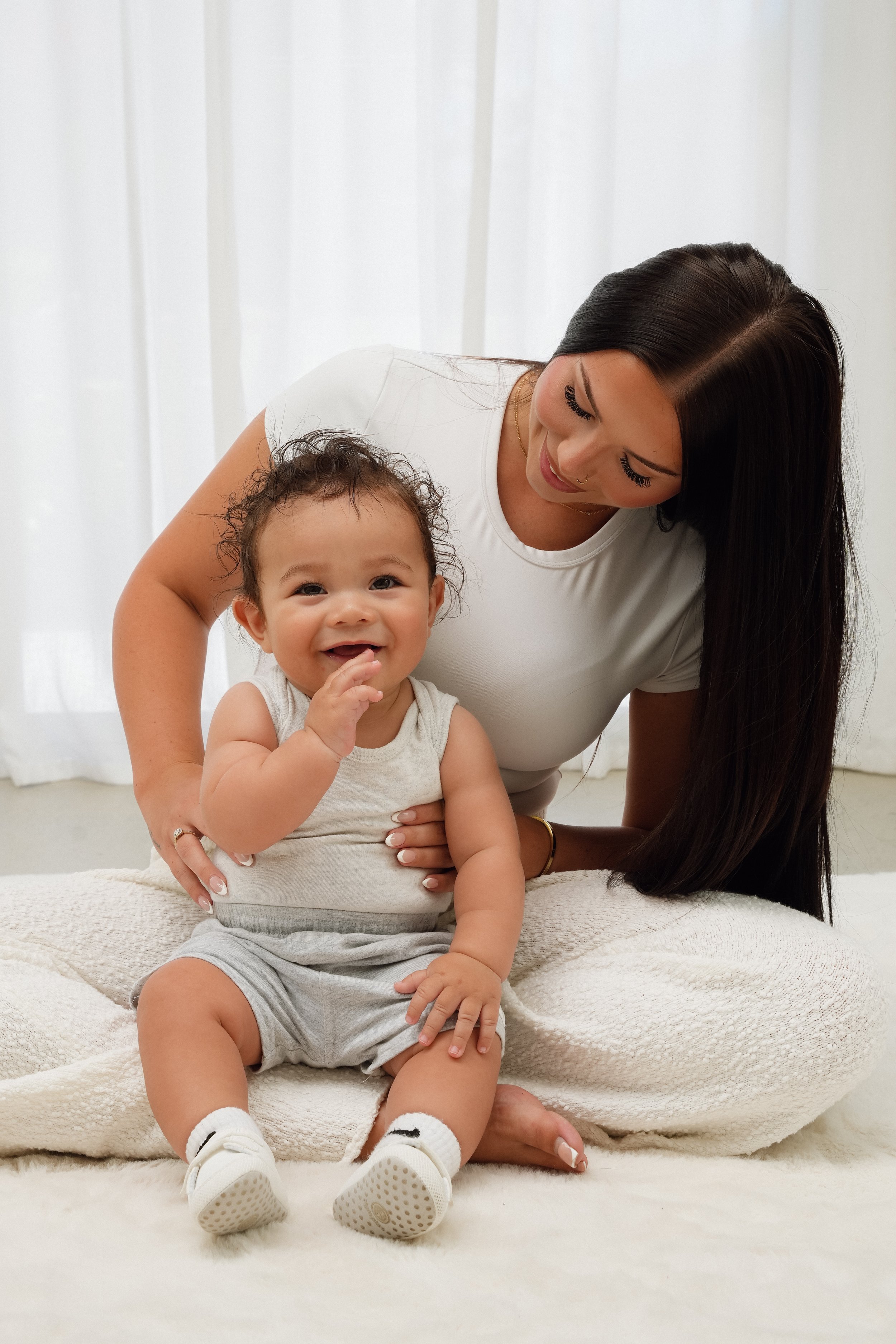 A woman and a baby sitting together on a white blanket in front of white curtains. The woman has long dark hair and is smiling at the baby, who is also smiling and touching his mouth.