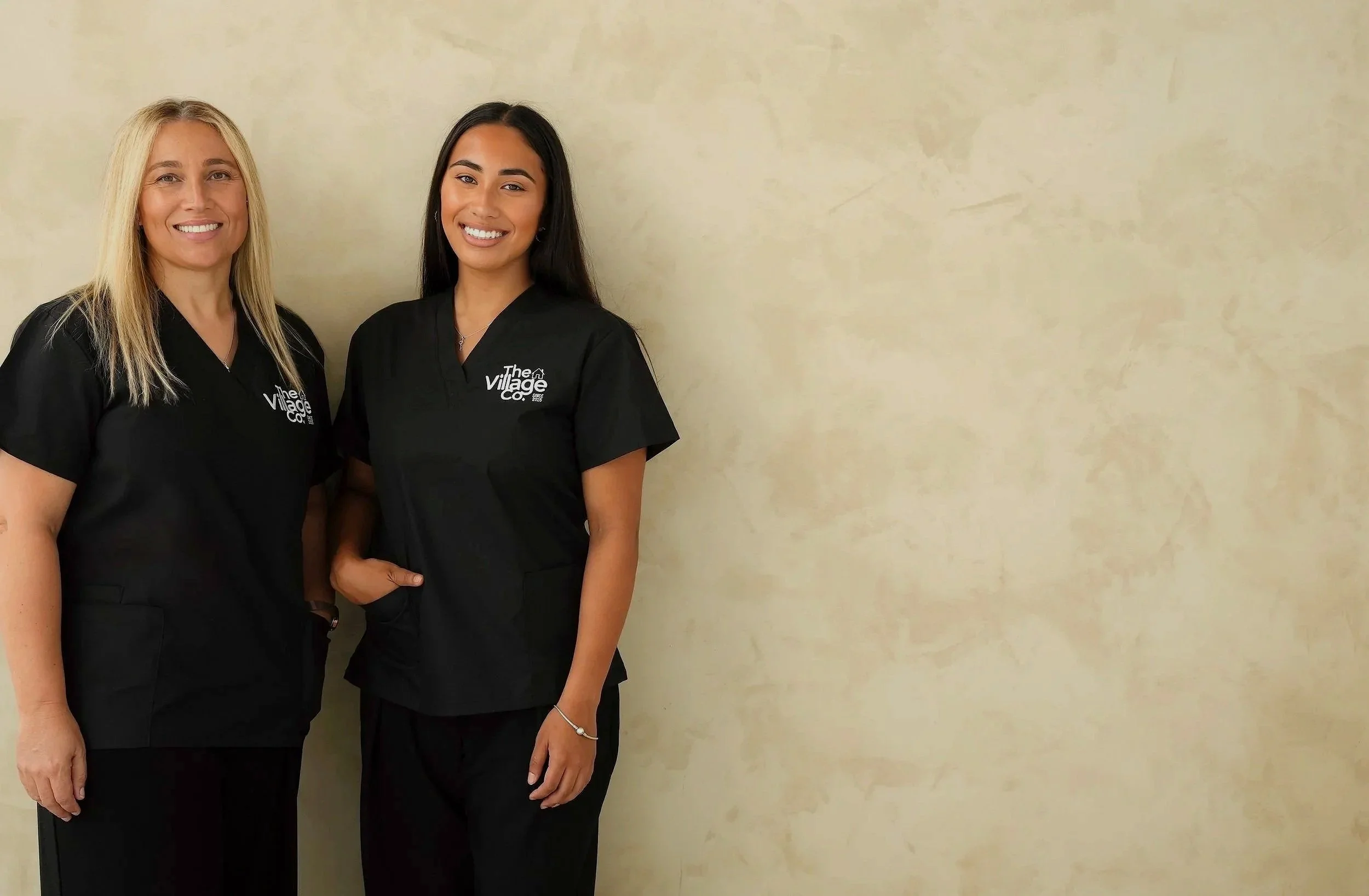 Two smiling women in black scrubs standing next to each other against a light beige wall, with no other objects in the image.