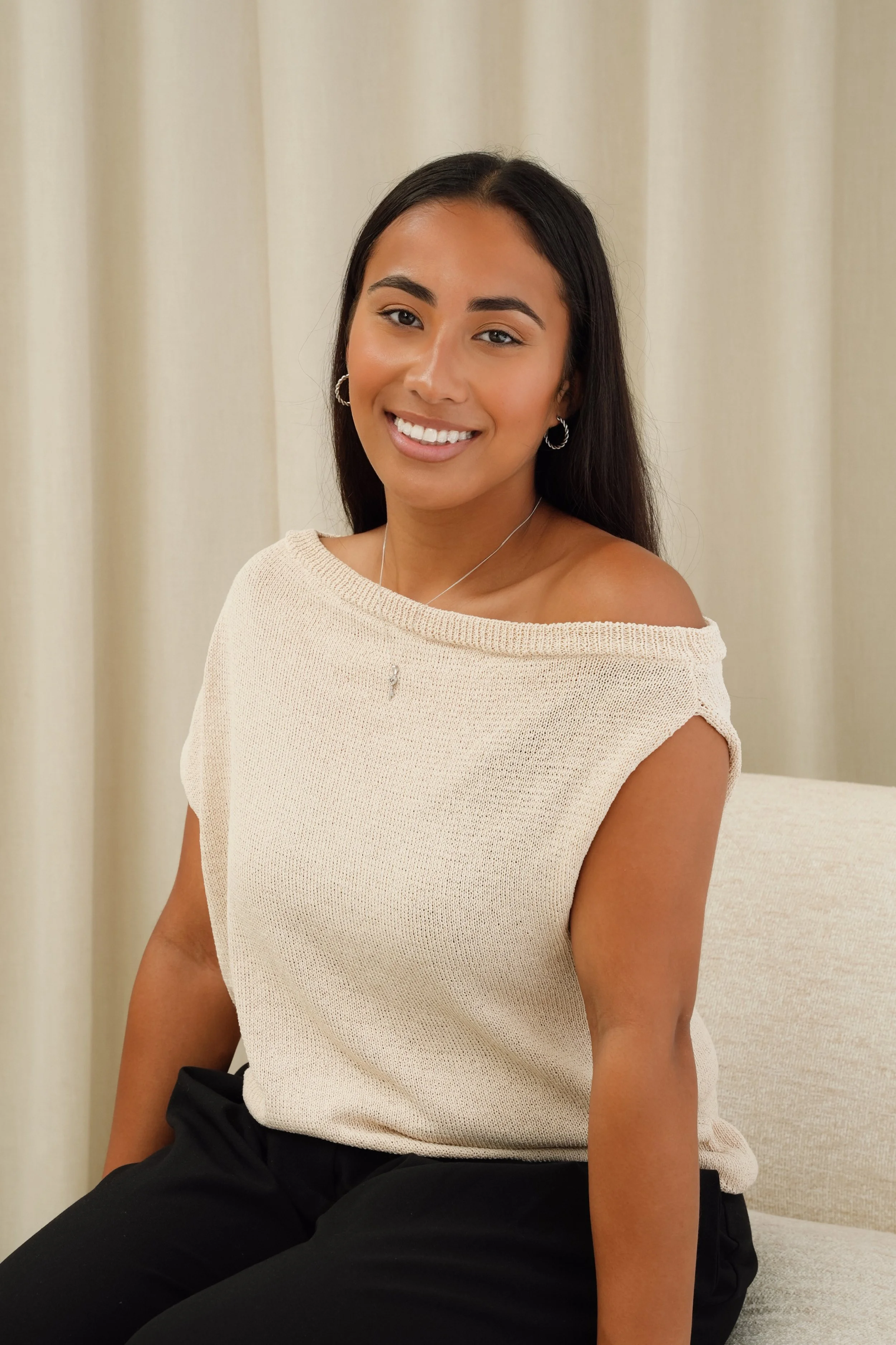 A young woman with long black hair, smiling, sitting on a beige couch in front of a cream-colored curtain, wearing a beige off-shoulder top and black pants.