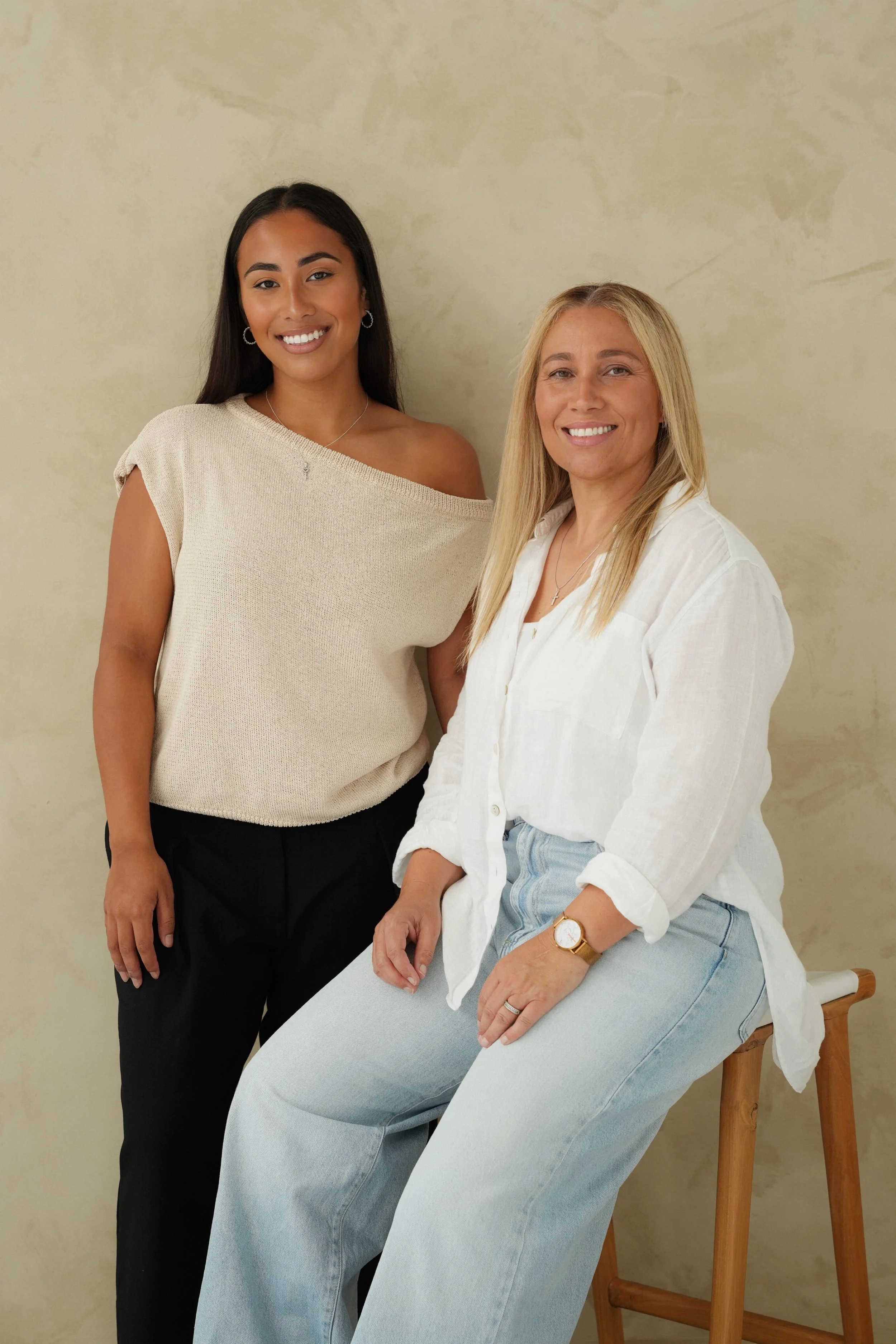 Two women smiling, one standing and one sitting on a wooden stool, in front of a beige textured background.
