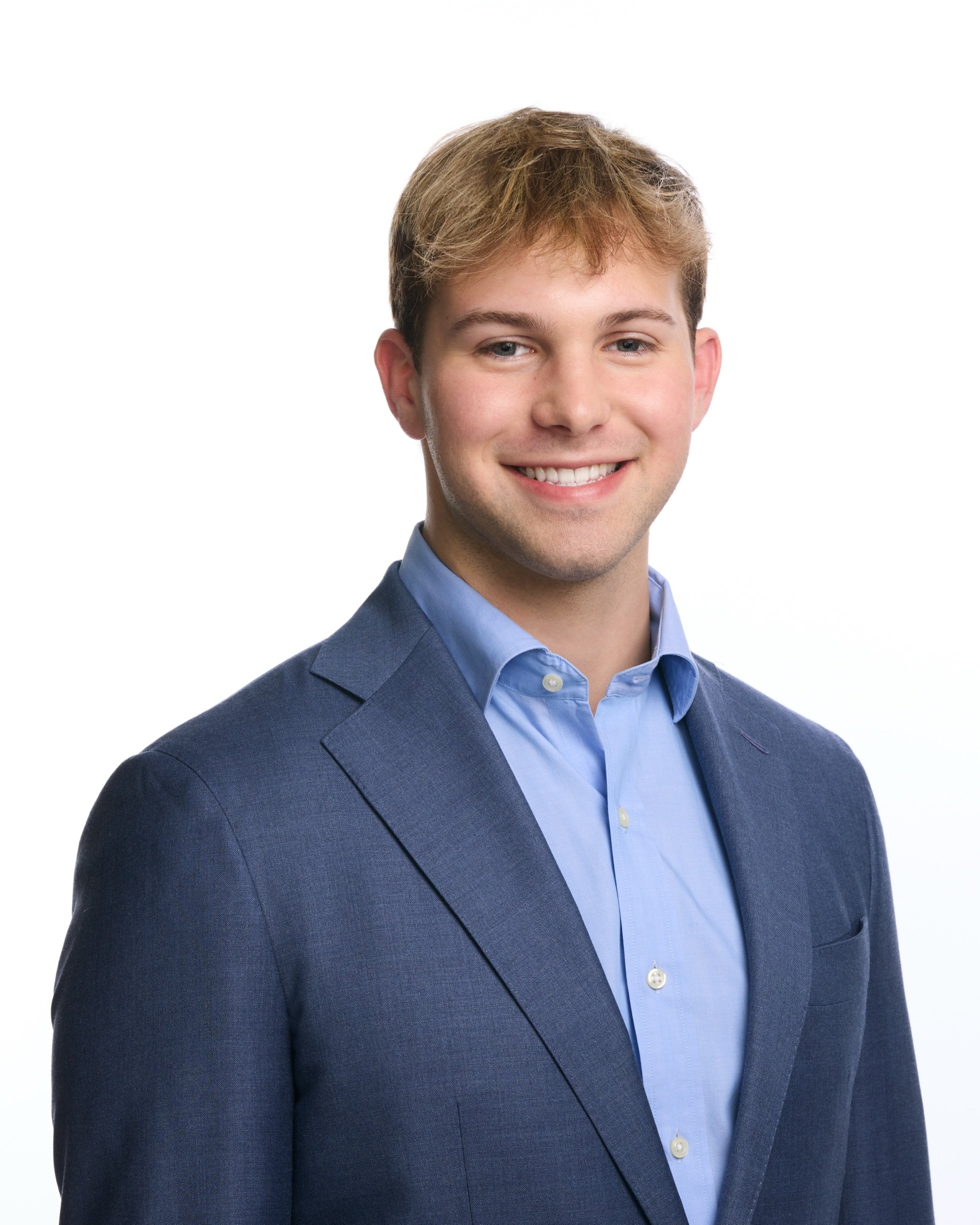 Young man wearing a blue suit and light blue shirt smiling against a white background.