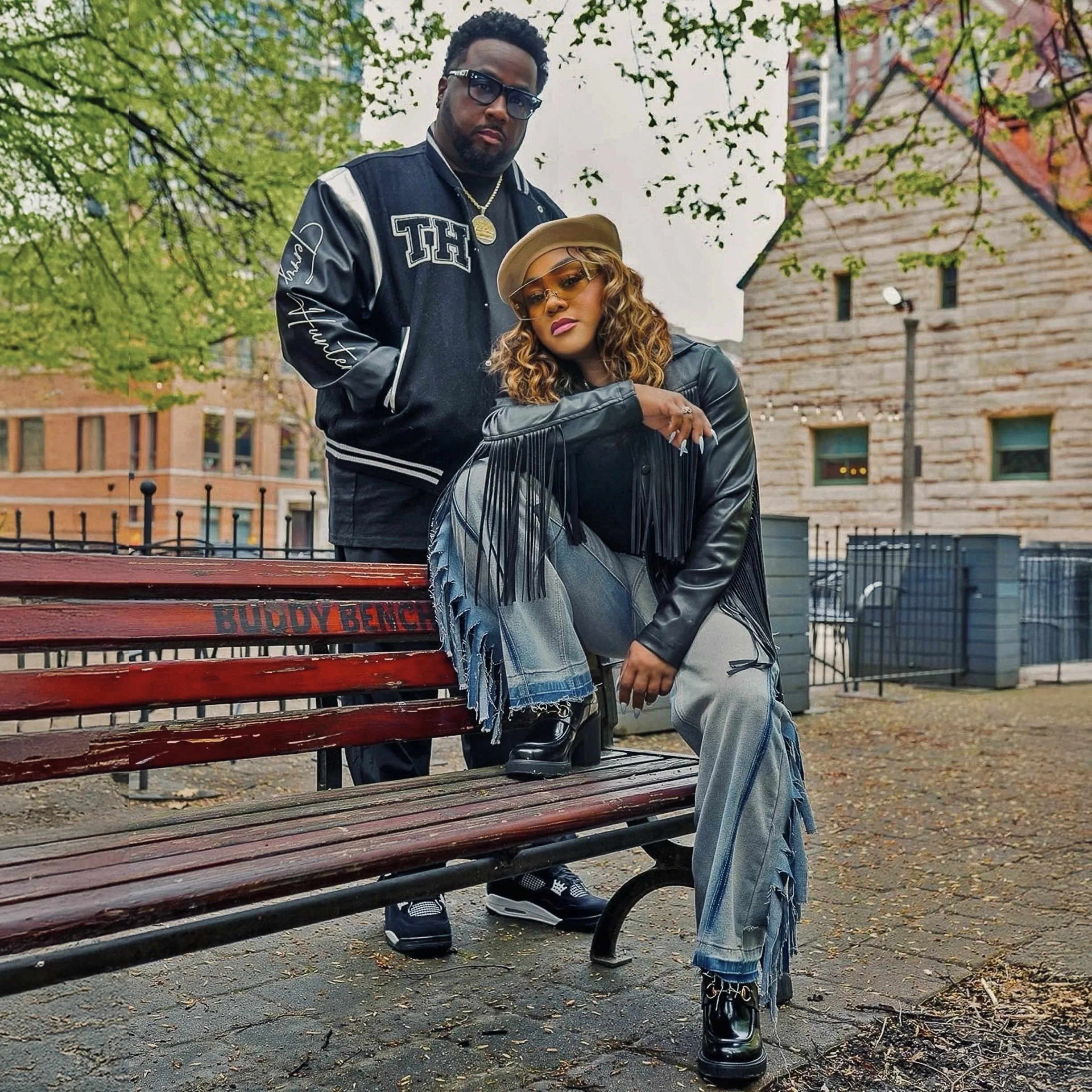 A man and a woman posing outdoors in a park with trees and buildings in the background. The woman is sitting on a park bench with one knee on the seat, wearing a black leather jacket with fringe, gray pants with fringe details, black boots, and sunglasses. The man is standing behind her, wearing a black jacket, glasses, and gold jewelry.