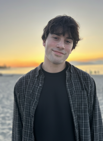 Young man with dark curly hair wearing a black T-shirt and open plaid shirt, standing on a beach at sunset.