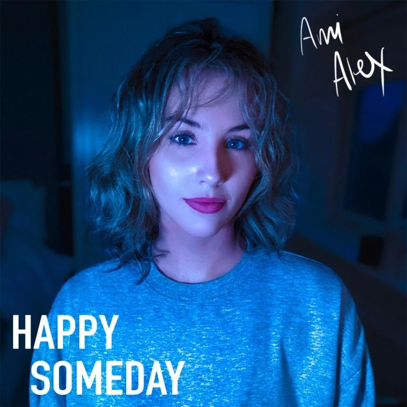 A young woman with wavy, shoulder-length hair and blue eyes taking a selfie in a dimly lit room with blue lighting, wearing a gray shirt. The image includes handwritten text "Am Alex" in the top right corner and large white text "HAPPY SOMEDAY" in th