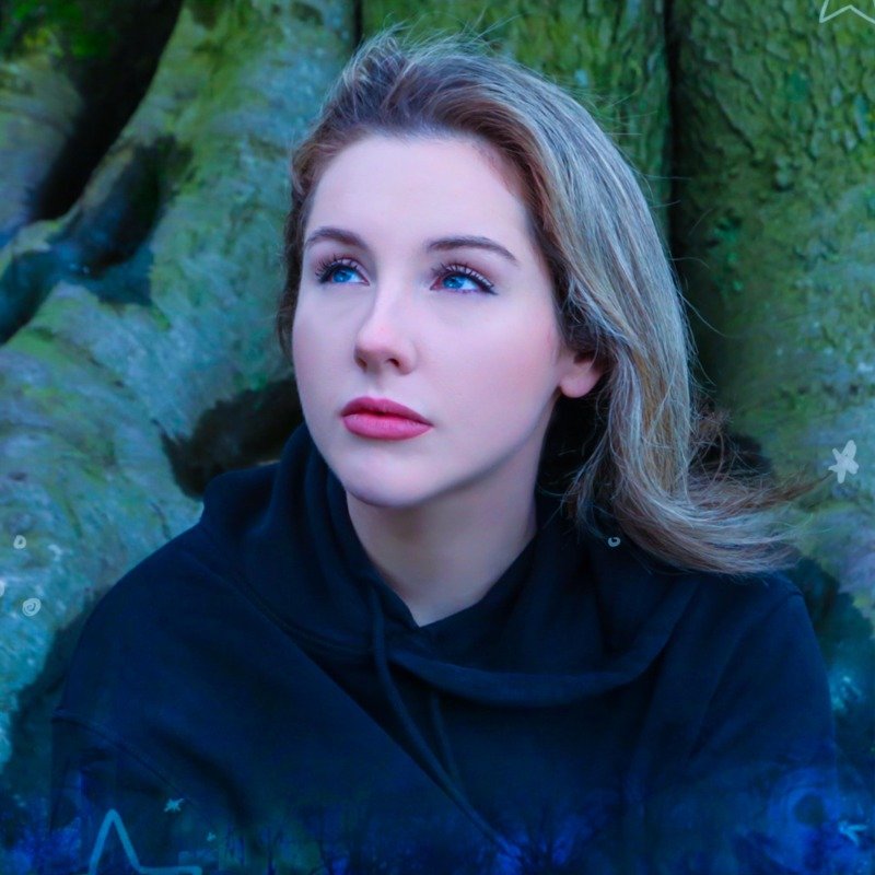 A young woman with wavy blonde hair and blue eyes looking upwards, wearing a dark hoodie, outdoors near a moss-covered rock formation.