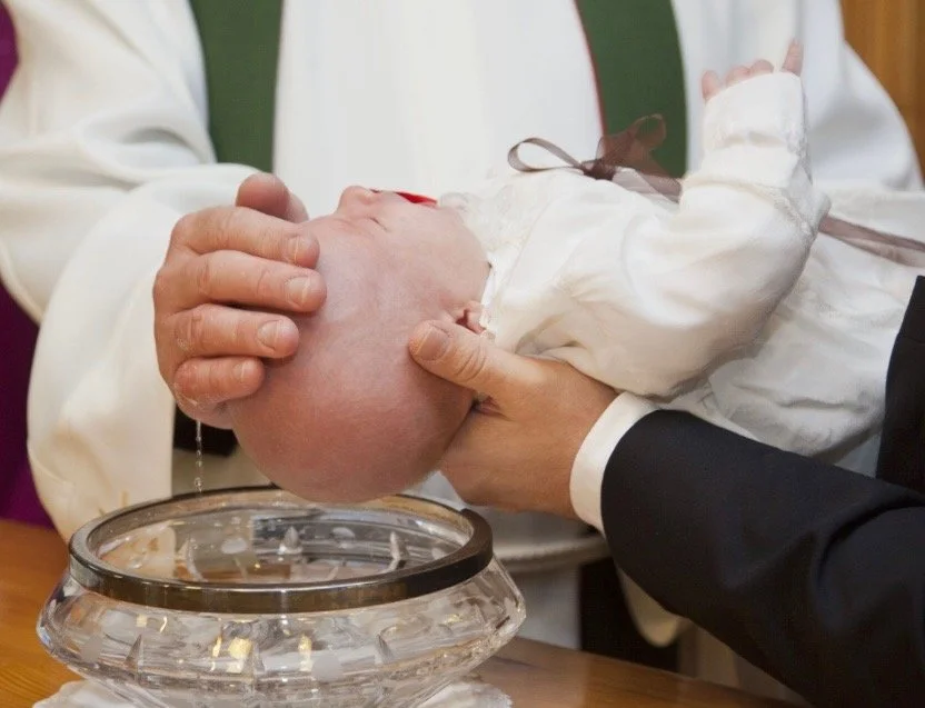A person in a white robe holds a baby's head over a glass baptismal font filled with water, during a baptism ceremony.