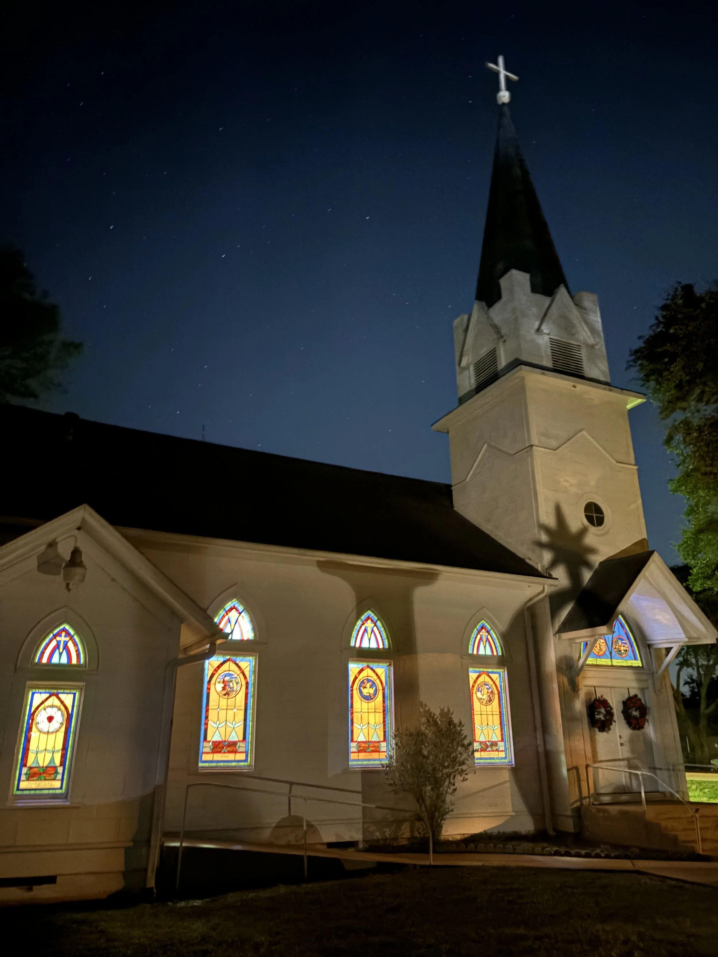 Night view of a small church with stained glass windows, a tall steeple with a cross, and a shadow of the cross cast on the church wall, under a starry sky.