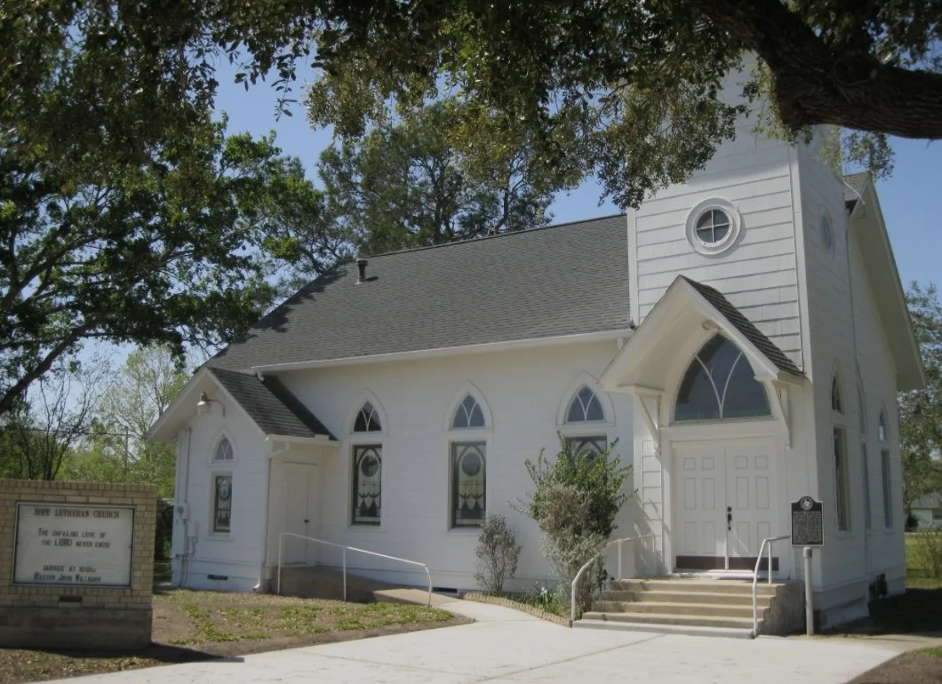 White church with a dark shingled roof, arched stained glass windows, a small steeple, and a sign near the entrance that reads 'New America Church'.