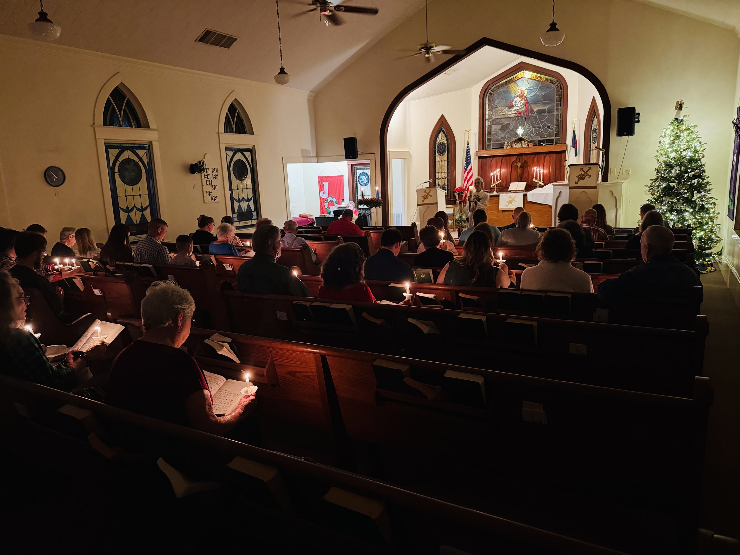 Inside a church sanctuary during a candlelight service, with people seated in pews, holding candles, and facing an altar with decorations including a Christmas tree, an American flag, and stained glass windows.