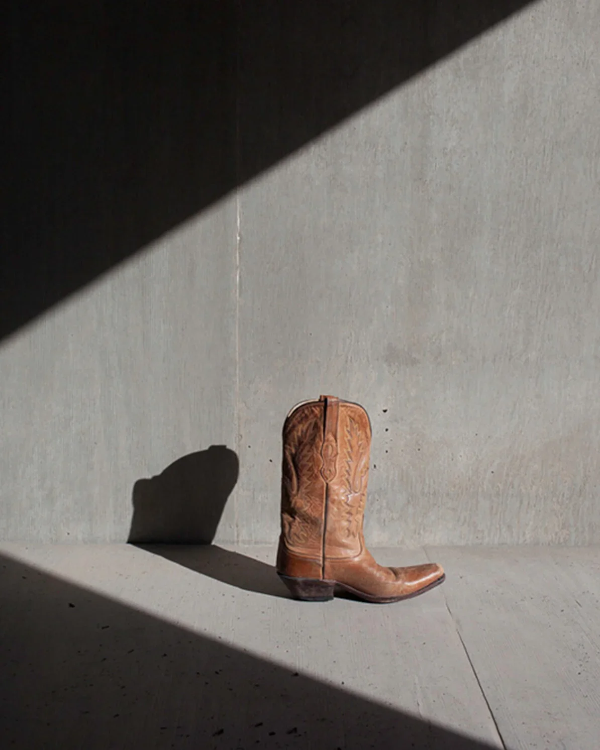 A brown leather cowboy boot standing alone against a light gray concrete wall, casting a shadow on the floor.