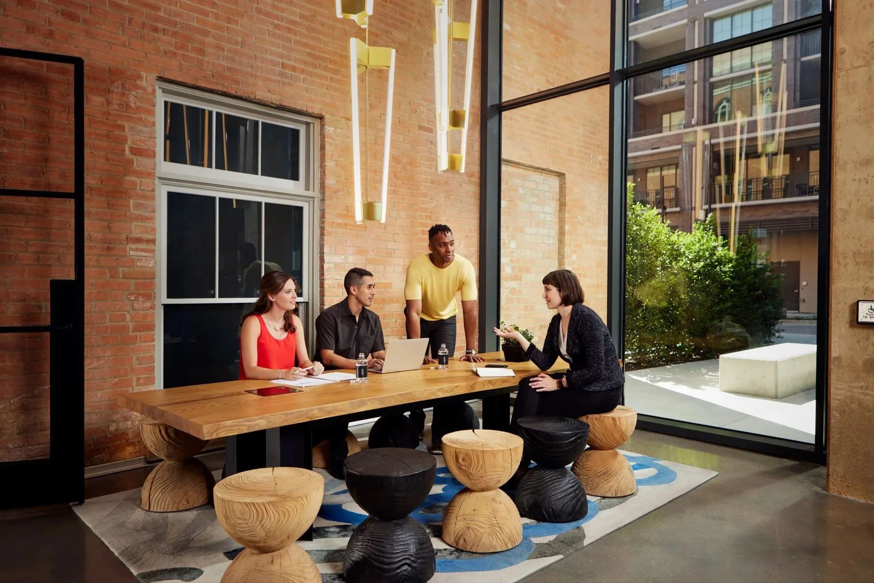 A group of four young professionals having a meeting in a modern conference room with large glass windows, brick walls, and contemporary hanging lights.