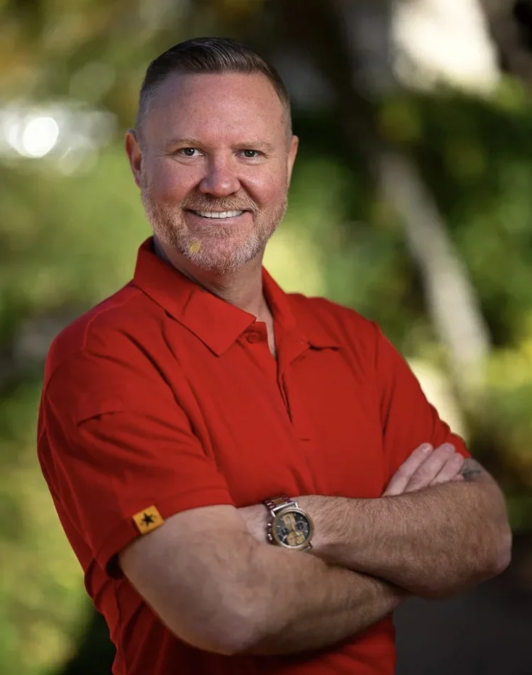 Smiling man with a beard and short hair, wearing a red collared shirt, arms crossed, outdoors with greenery background.