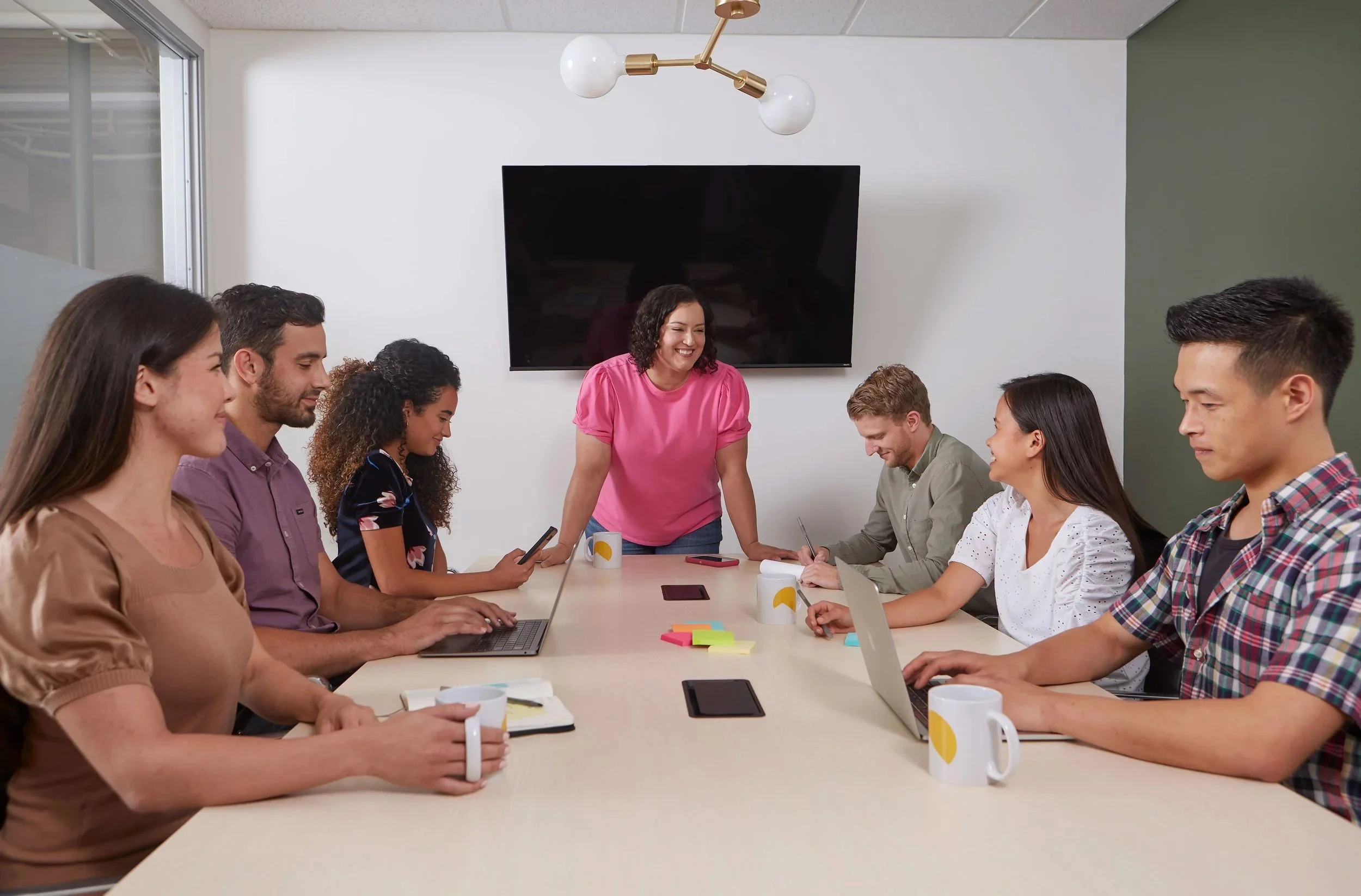 Group of people sitting around a conference table, engaged in a meeting with a woman standing and smiling at the center of the room, with laptops, coffee mugs, notebooks, and colorful sticky notes on the table, in a modern office environment.