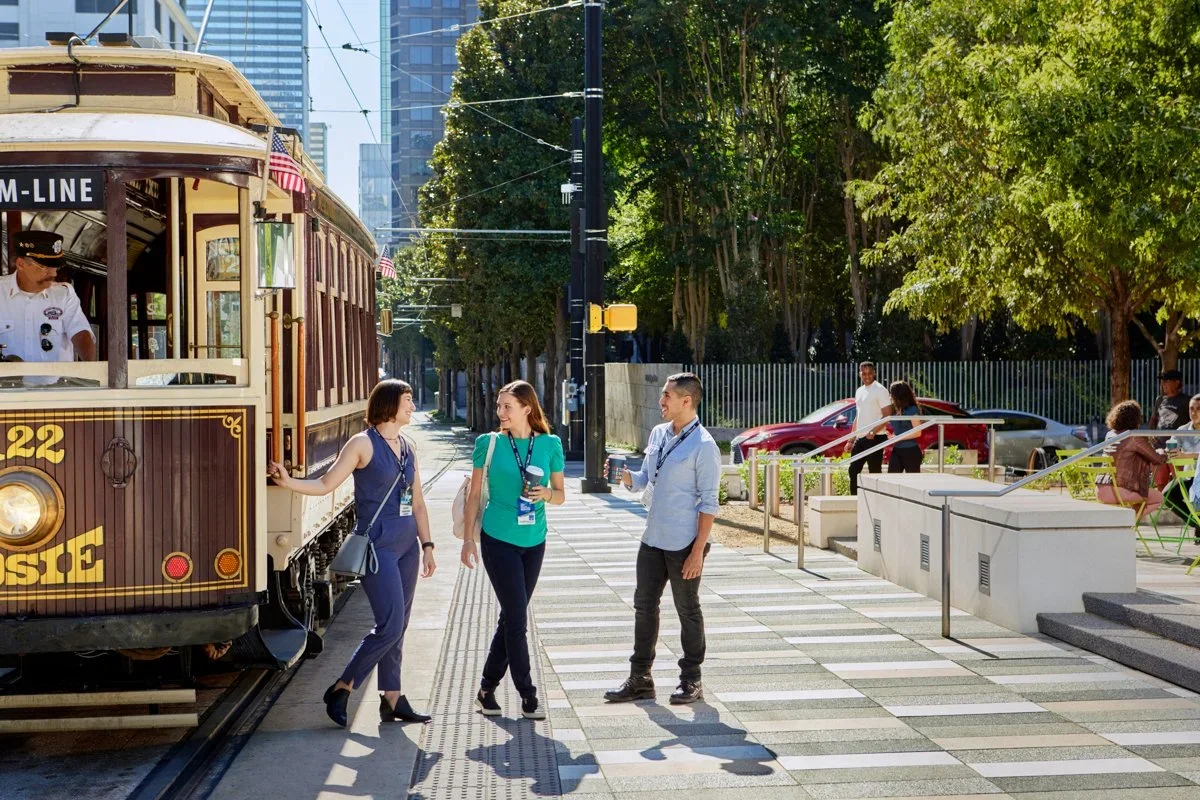 Three people are standing on a city sidewalk next to a vintage trolley car, engaging in conversation. The trolley is brown with cream trim, and a conductor is visible inside. There are trees and parked cars in the background, with other pedestrians s