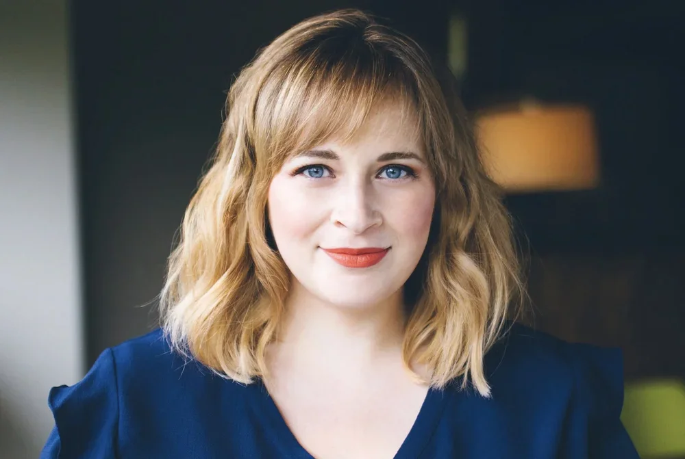 A woman with shoulder-length wavy blonde hair, blue eyes, and wearing a blue top, smiling in an indoor setting with a blurred background.