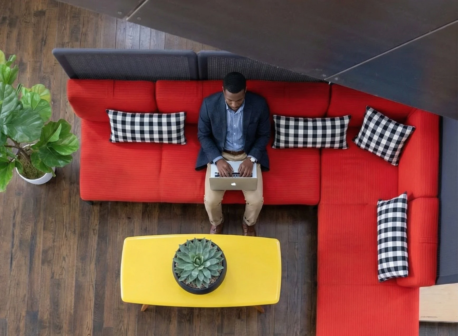 A man sitting on a red sectional sofa working on a laptop, surrounded by checkered pillows, with a yellow table holding a succulent plant and a potted green plant nearby.