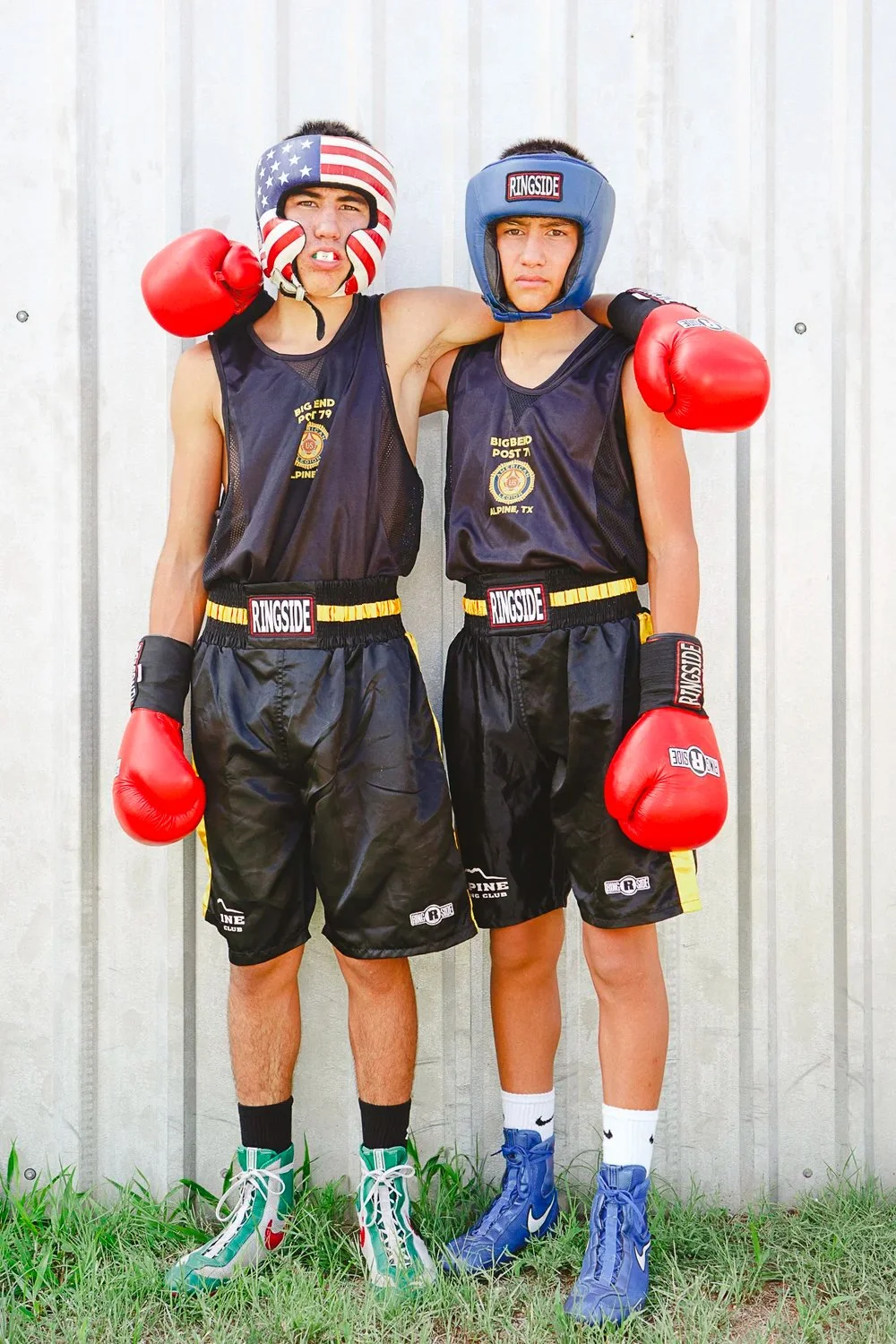 Two young men in boxing gear standing side by side outdoors against a metal wall, with arms around each other. Both wear black boxing uniforms with red boxing gloves. The man on the left wears a headband with an American flag pattern, and the man on 