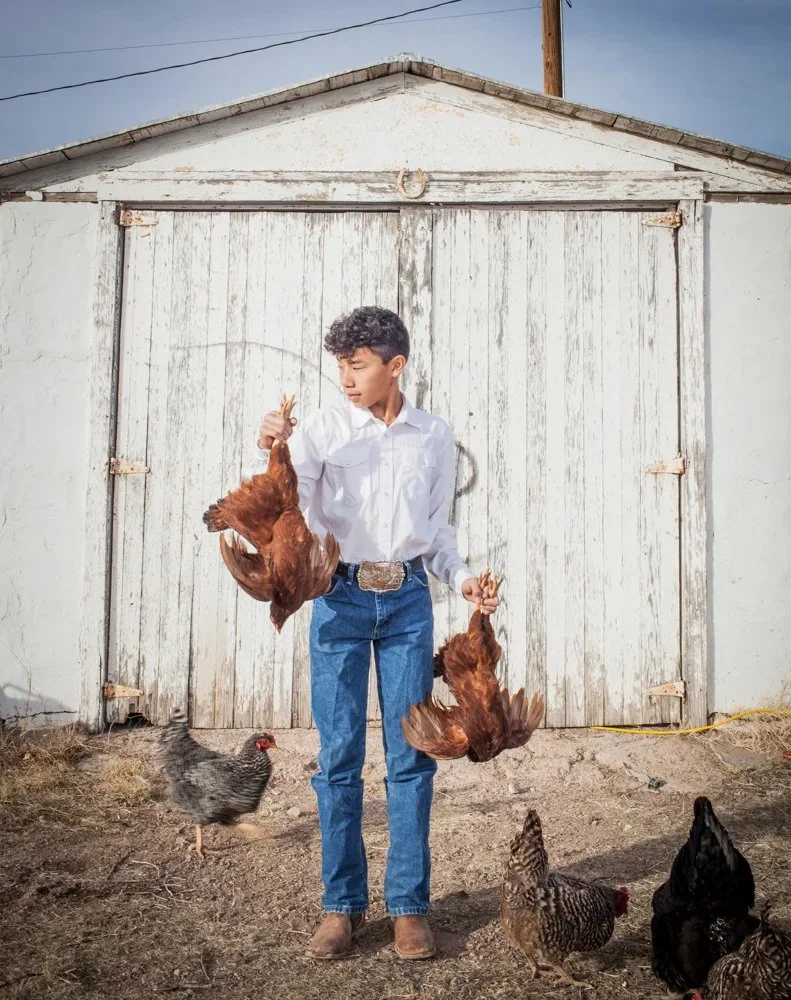 A young man with curly hair wearing a white shirt, blue jeans, and brown boots stands outside in front of a white wooden barn door, holding two brown chickens, one in each hand. Several other chickens, including a black one and a speckled one, walk o