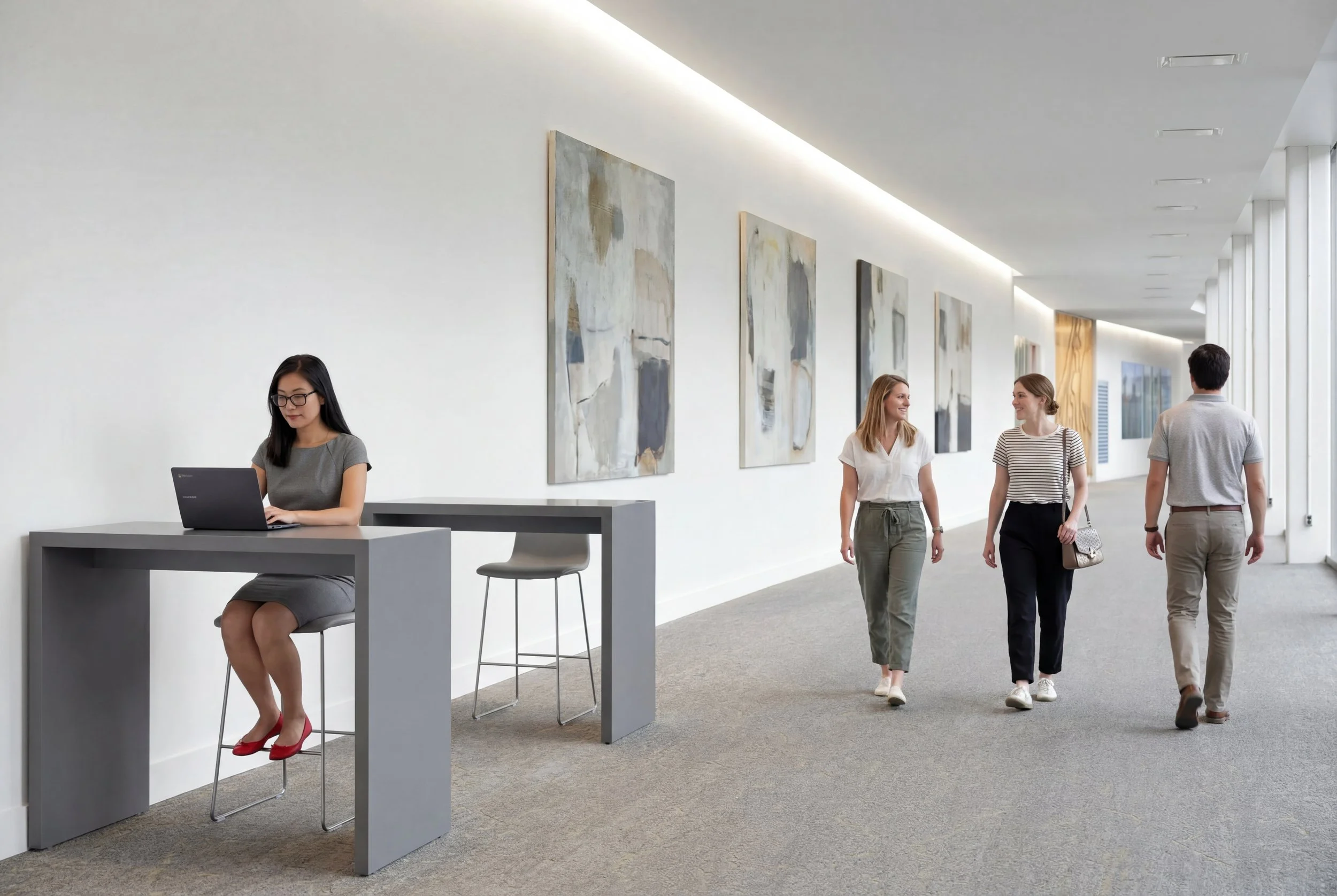 A woman in a gray dress sitting at a high desk working on a laptop in a modern art gallery with abstract paintings on the wall, while three people walk and talk in the background.