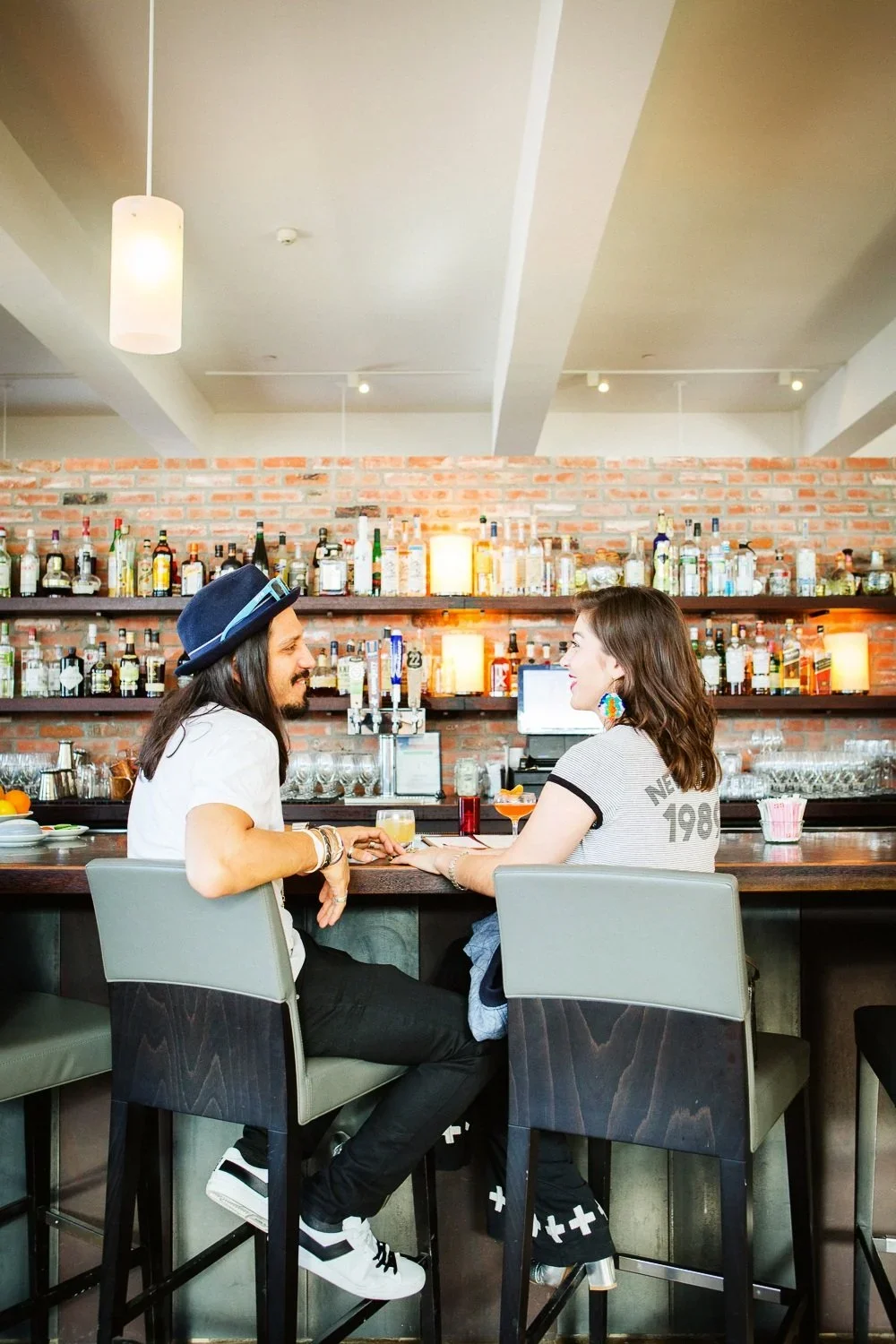 A man and woman sitting at a bar, holding hands and smiling at each other, with alcohol bottles on shelves behind them.