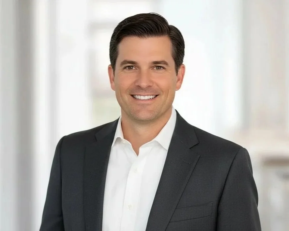 A man wearing a dark suit and white shirt smiling at the camera, standing in a bright, modern office with blurred background.