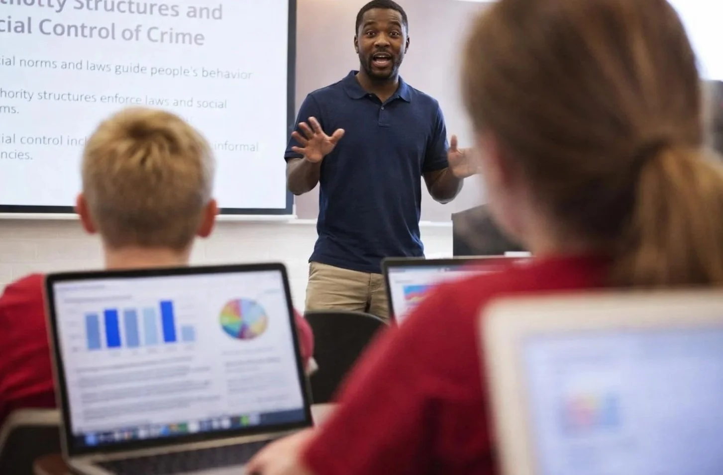 A teacher or presenter speaking to students in a classroom while using a projector screen. The students are using laptops with graphs and charts displayed.