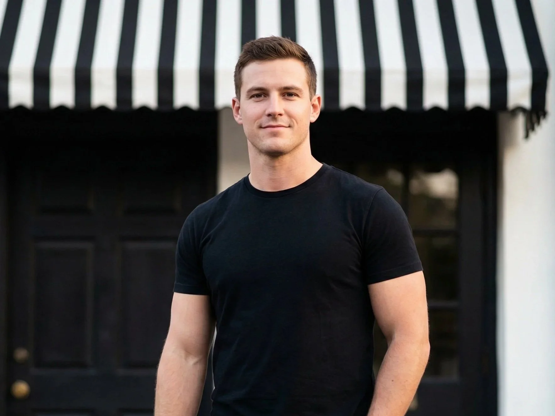 A young man with light skin, short brown hair, wearing a black T-shirt, standing outdoors in front of a building with black double doors and a black and white striped awning.