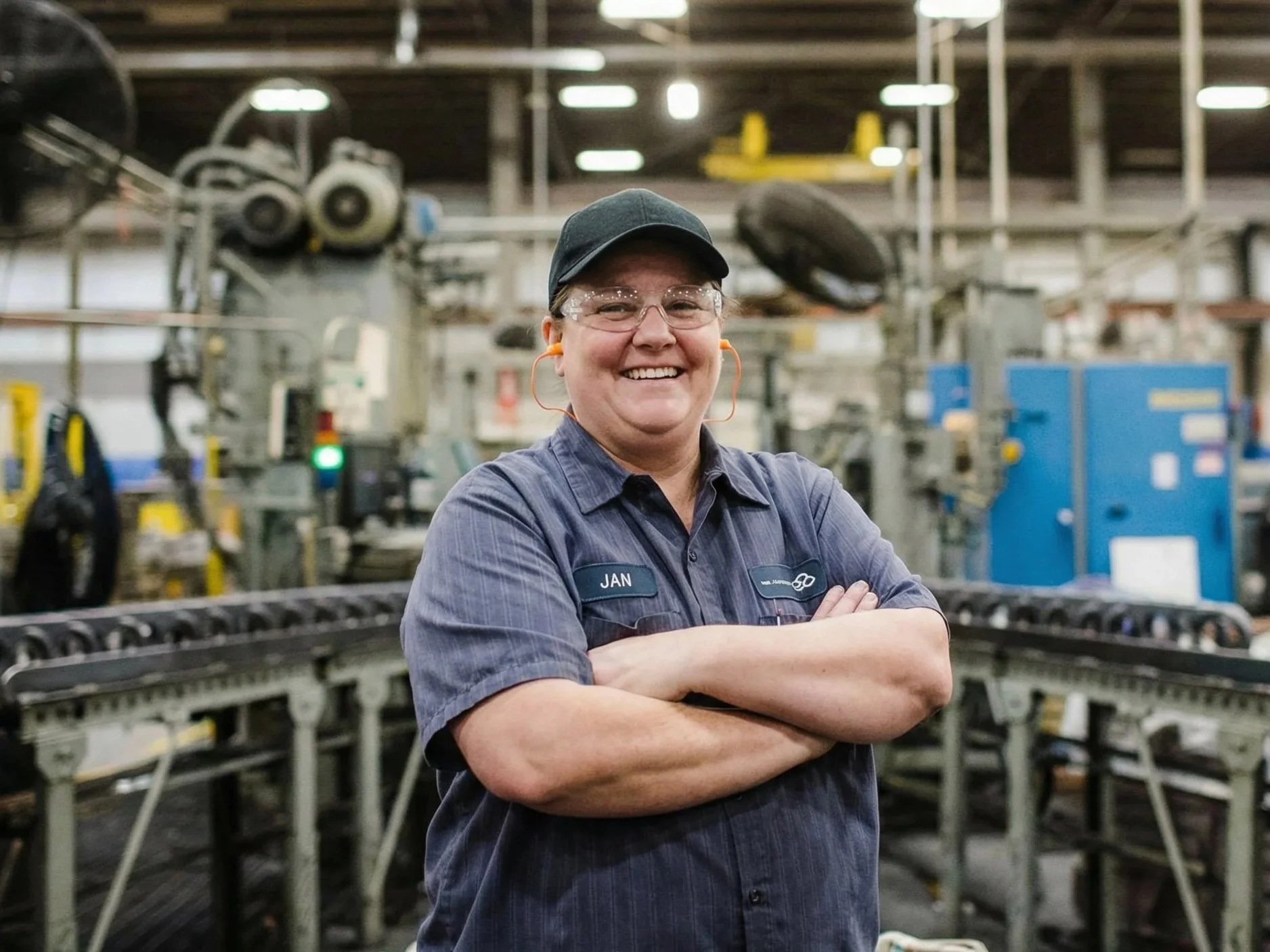 A smiling woman in work uniform standing with arms crossed in an industrial factory setting with machinery in the background.