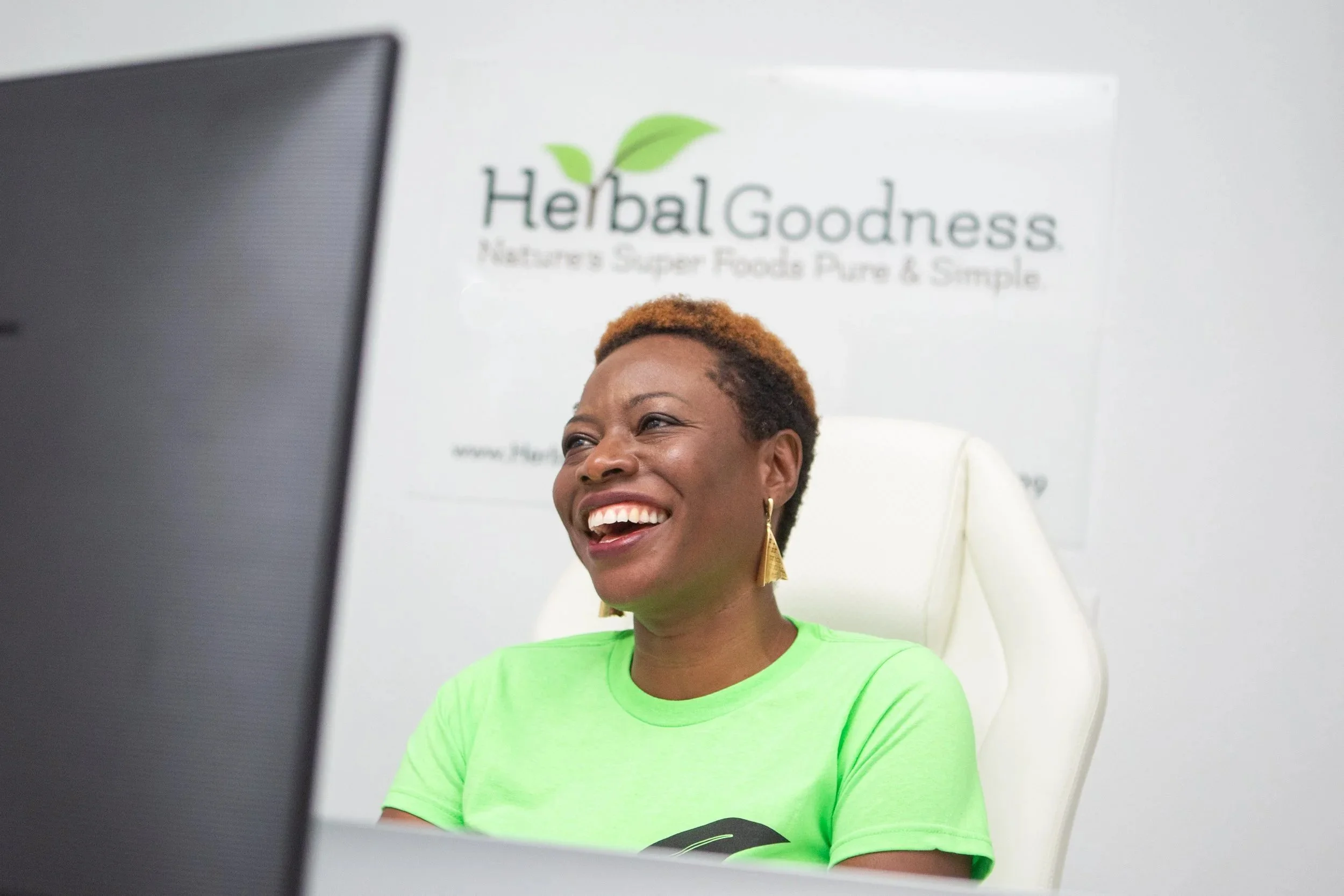 Smiling woman sitting at a desk with a computer monitor in front of her, in an office with a sign that says 'Herbal Goodness' in the background.