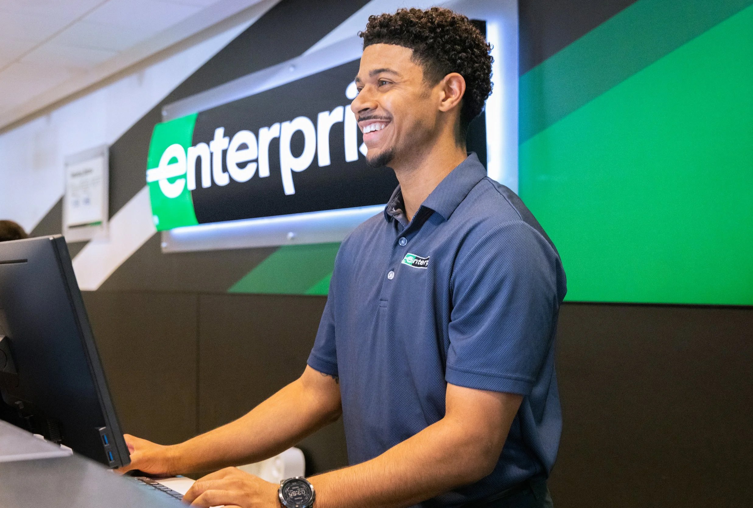A smiling man wearing a navy blue polo shirt standing at a counter with a computer, in front of a sign with the word 'enterprise' on a green and black background.