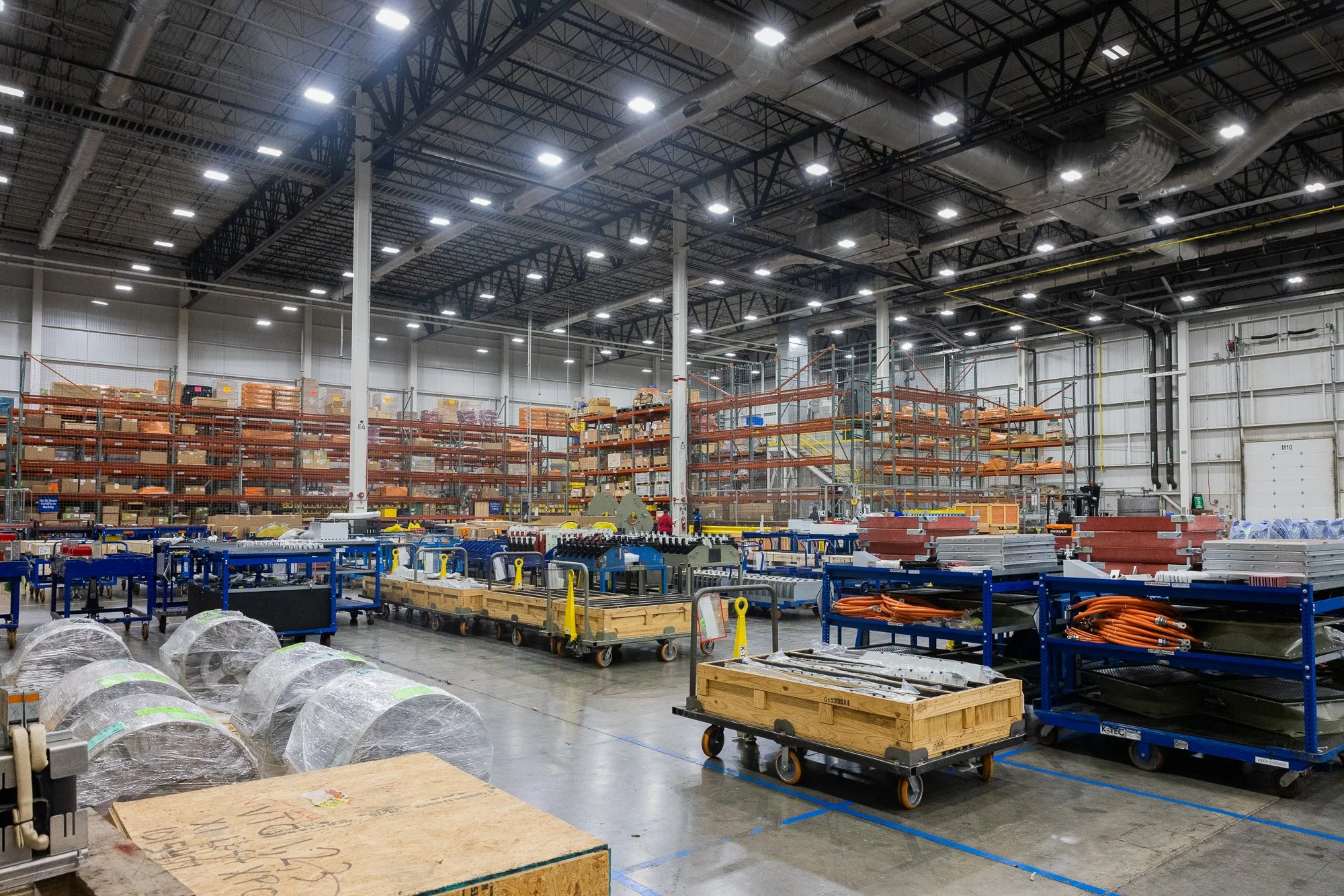 Interior of a large warehouse in Fort Worth with metal shelving filled with boxes and supplies, on a clean concrete floor with carts and wrapped rolls in the foreground, overhead lighting, and a large open space.