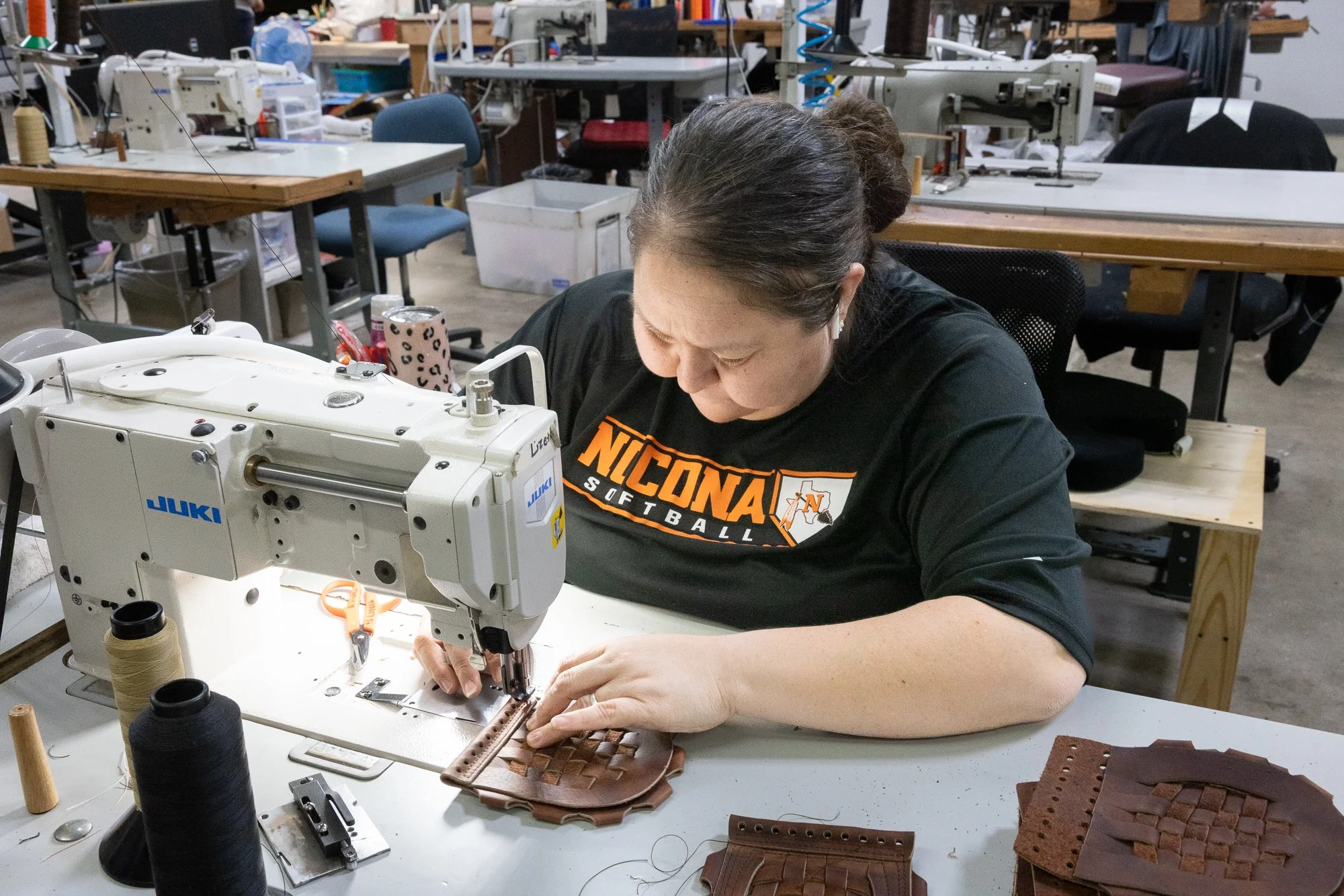 A woman using a sewing machine to work on leather woven material in a workshop or factory setting.