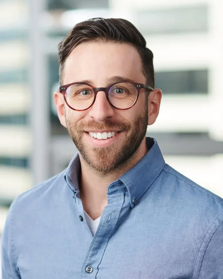 A smiling man with short dark hair and a beard, wearing glasses and a blue button-up shirt, standing in front of a blurred office building background.