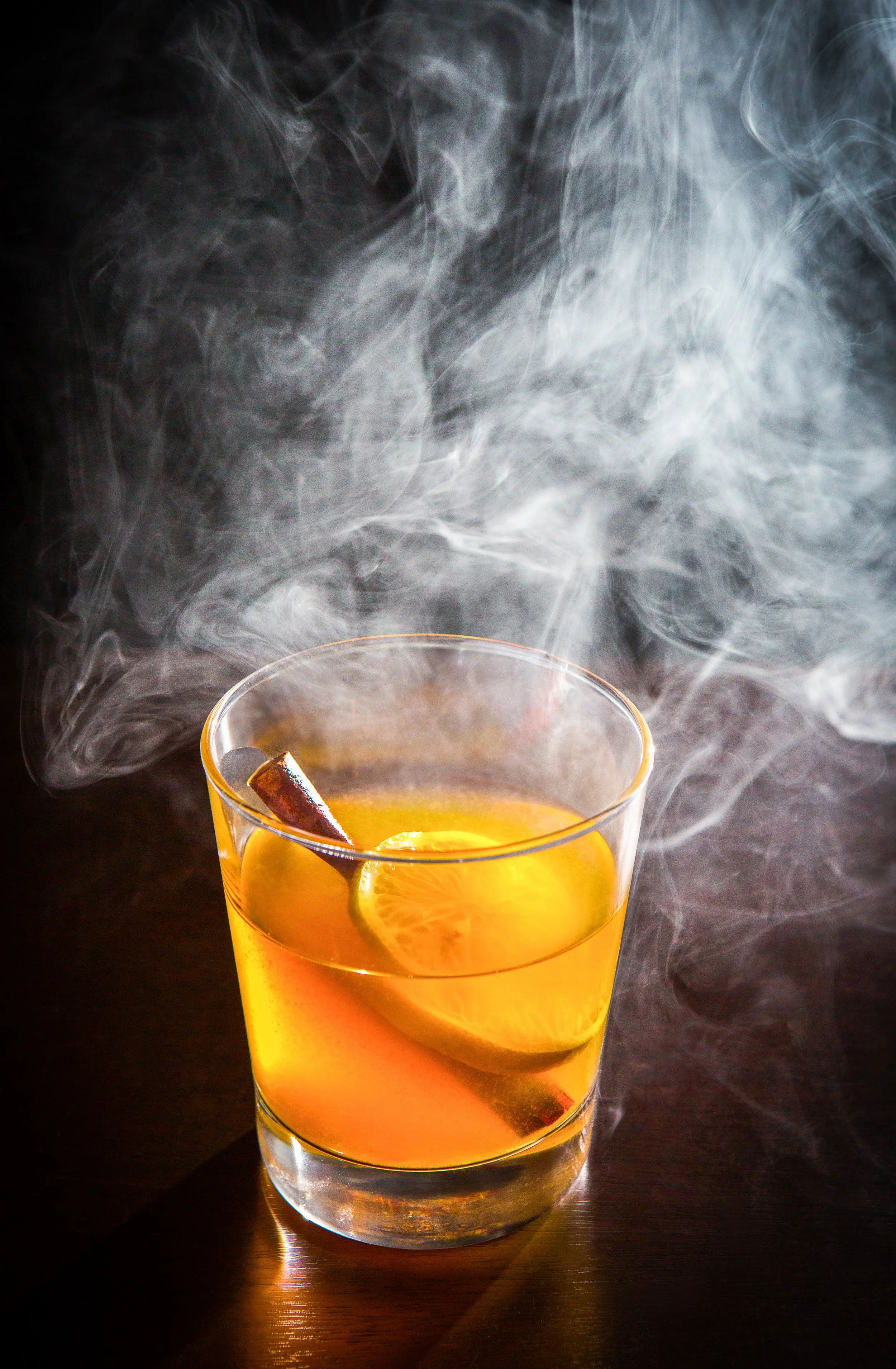 A glass of hot beverage with lemon slices and a cinnamon stick, emitting steam in a dark background.