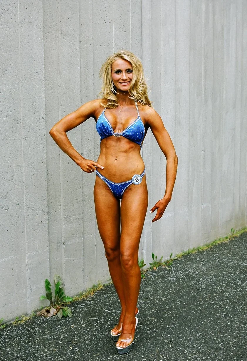 A woman with blonde hair wearing a blue bikini with rhinestone embellishments, standing against a concrete wall, smiling, with a contestant number badge on her bikini bottom.