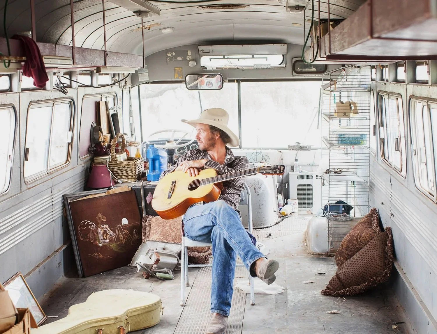 A man wearing a wide-brimmed hat and casual clothing sitting in a vintage bus, playing an acoustic guitar. The bus interior is cluttered with framed pictures, blankets, and various items, with large windows on sides and some shelves above.