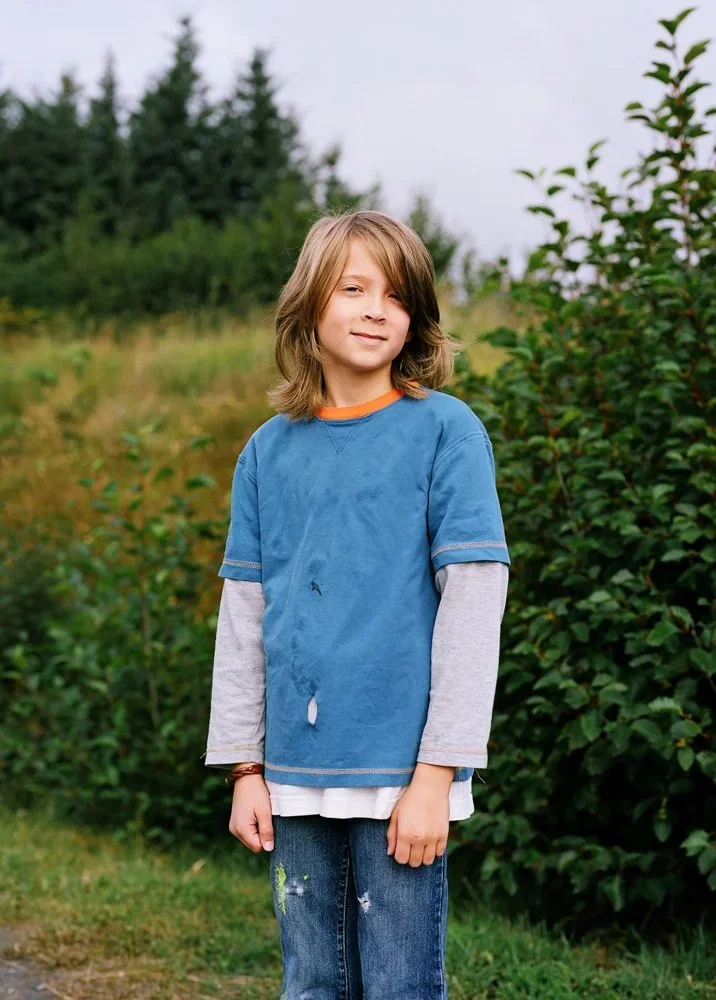 A young boy with light brown hair, wearing a blue t-shirt over a gray long-sleeve shirt and jeans, standing outdoors near green bushes and trees.