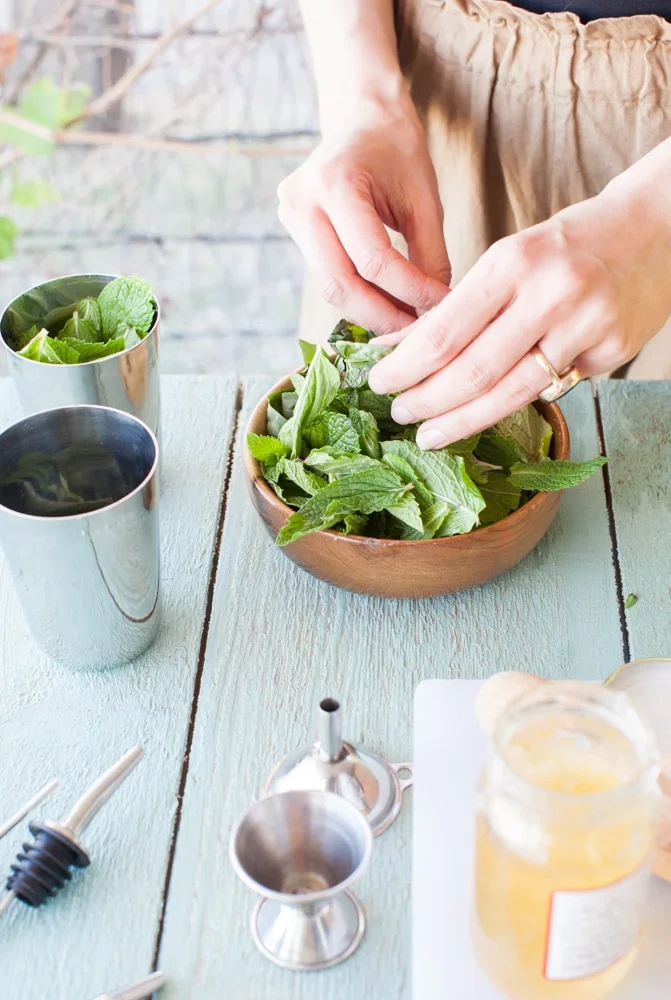 Person placing fresh mint leaves into a wooden bowl on a light blue table, surrounded by metal cups, a honey jar, and cocktail-making tools.