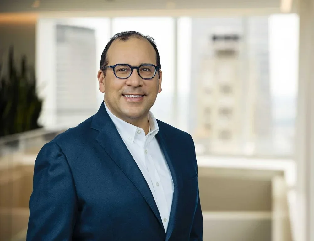 A man in a blue suit and glasses smiling in an office setting with large windows in the background.